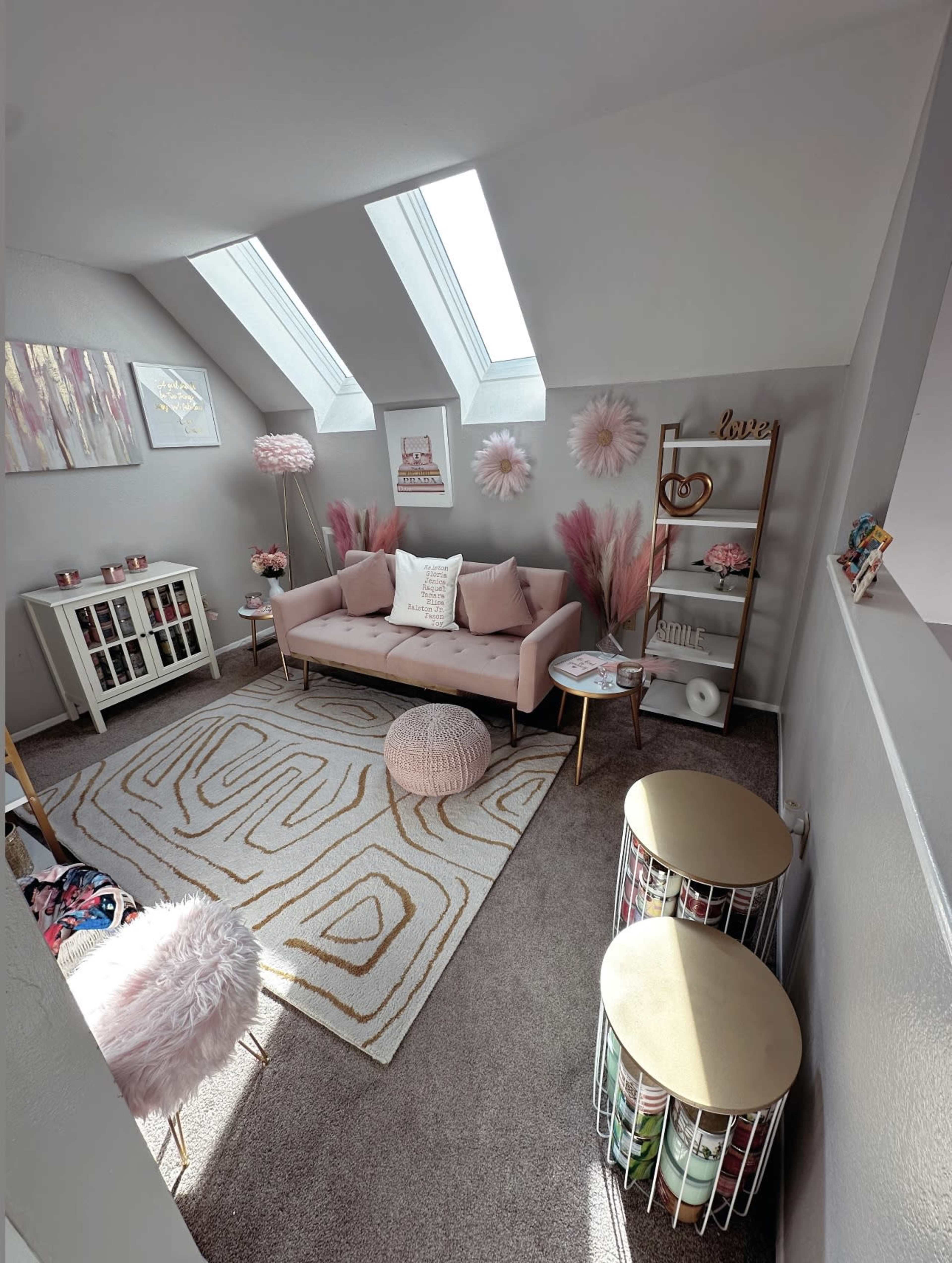 The image shows a cozy attic room with a pink sofa, decorative shelving, a patterned rug, and large skylights allowing natural light.