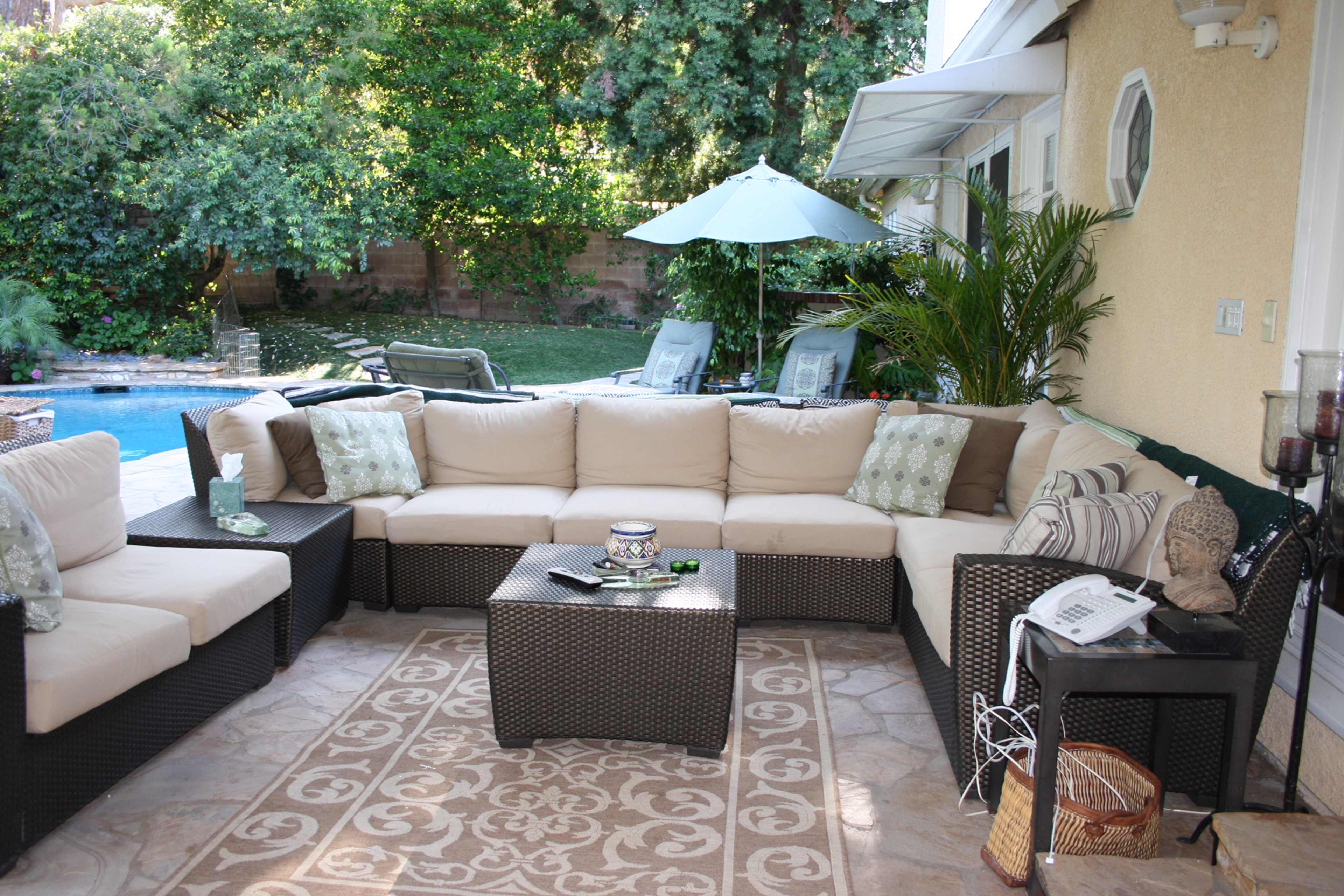 A covered outdoor seating area with a sectional sofa, a coffee table, and a backdrop of a swimming pool and green foliage.