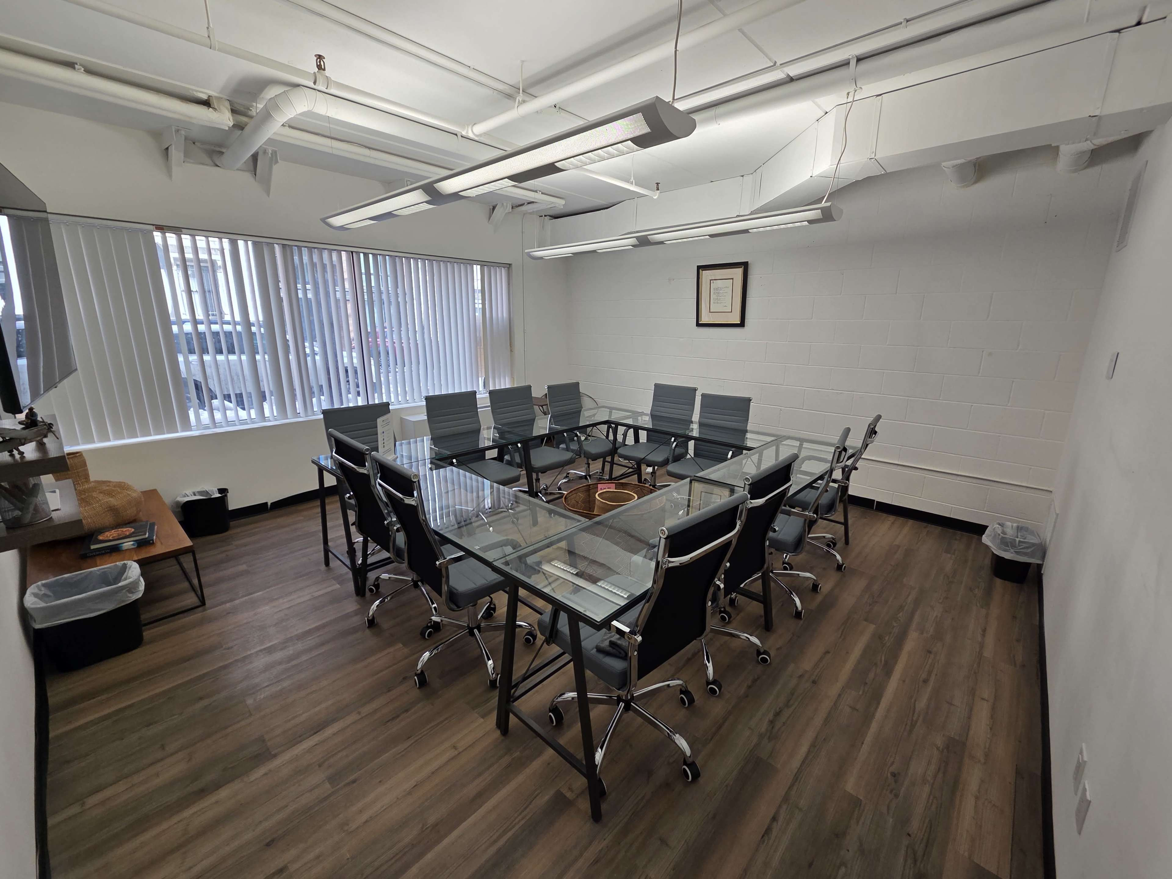 The image shows a modern conference room with a rectangular arrangement of glass-topped tables and gray office chairs, featuring large windows that allow natural light.