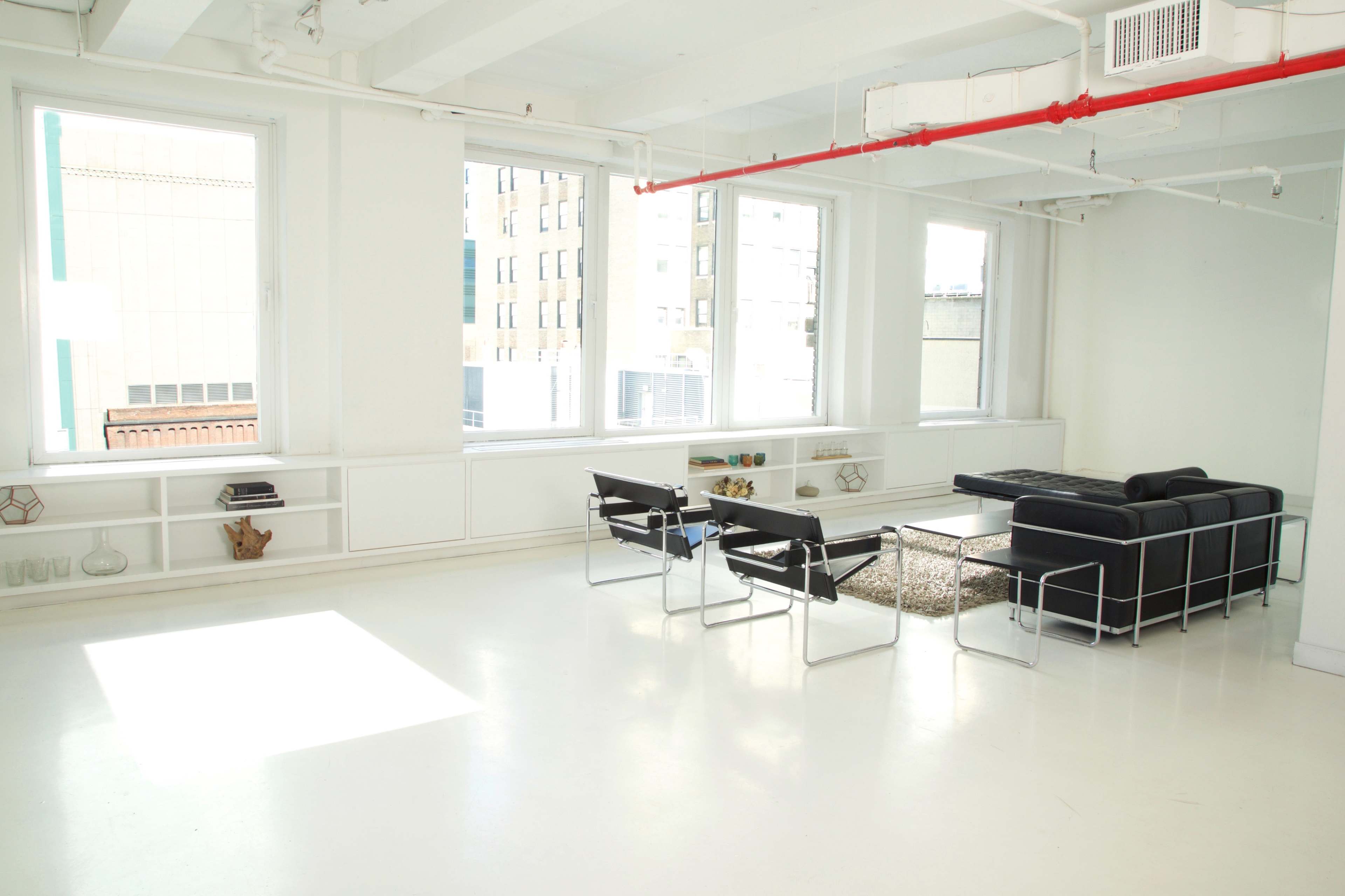 A spacious, minimalist interior with large windows, a black leather sofa, metal chairs, and a coffee table on a light-colored floor.
