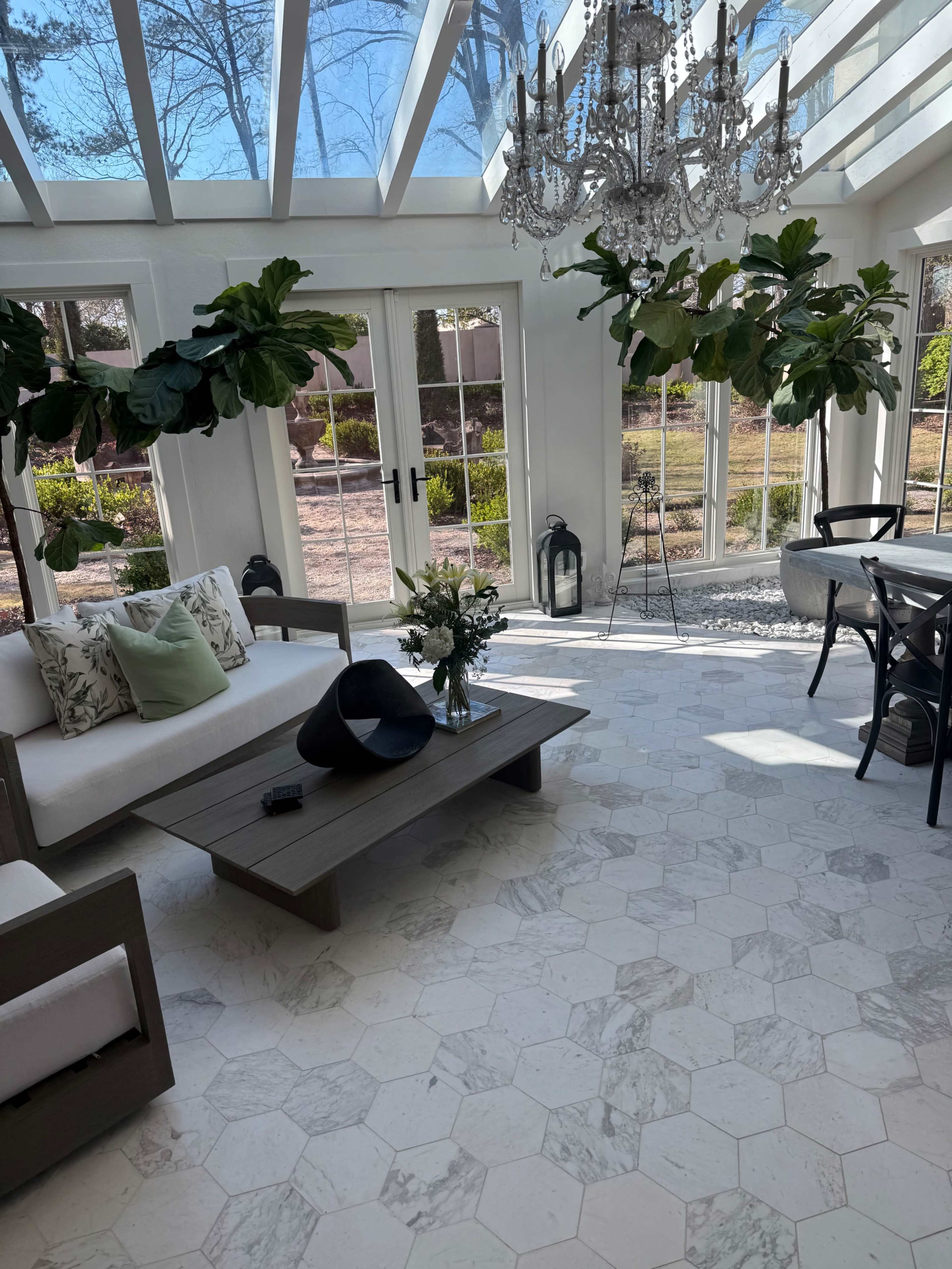 The image shows a bright sunroom with a chandelier, a large hexagonal marble floor, a coffee table, and potted plants hanging from the ceiling.