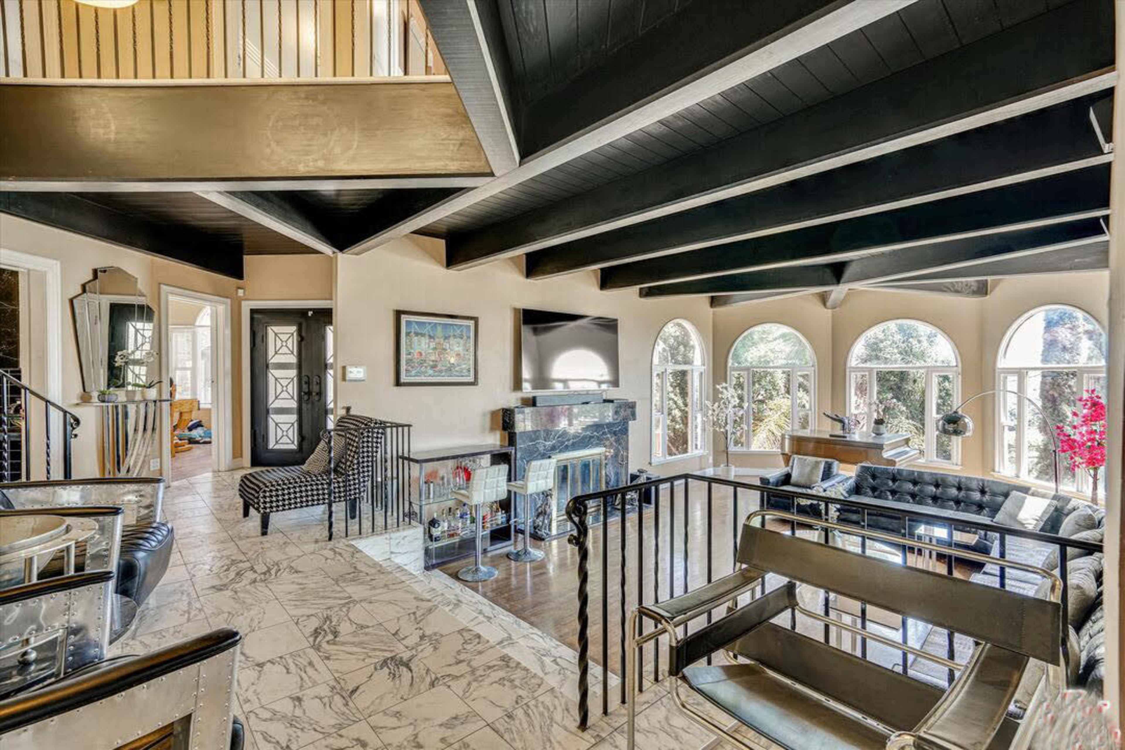 The image shows a spacious, modern living room with a high ceiling, large windows, and a mix of contemporary furniture.