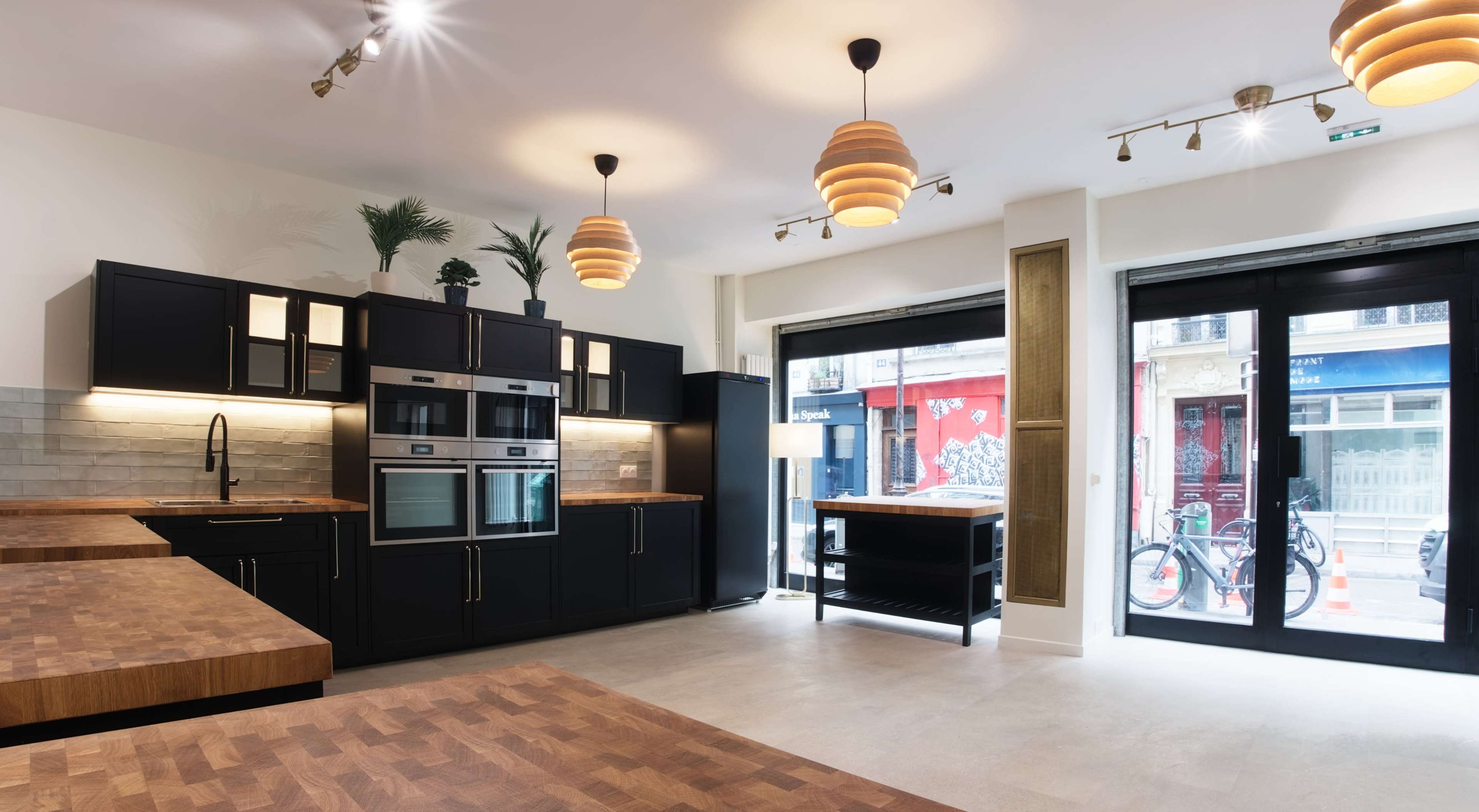 A modern kitchen features dark cabinets, a wooden countertop, and large windows overlooking a street with parked bicycles.