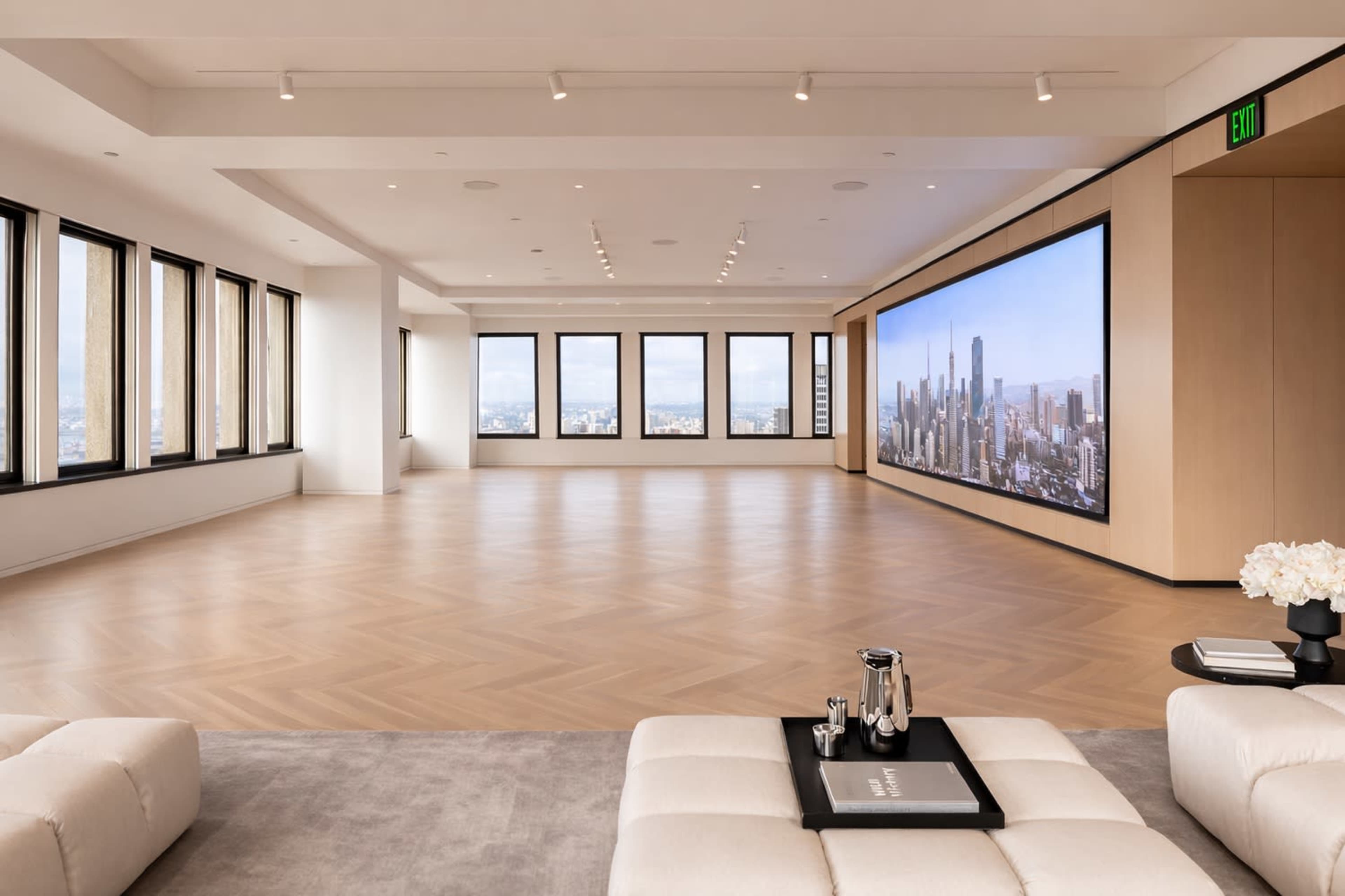 A large, modern room features a panoramic view of a city skyline through multiple windows and a large digital display on one wall.