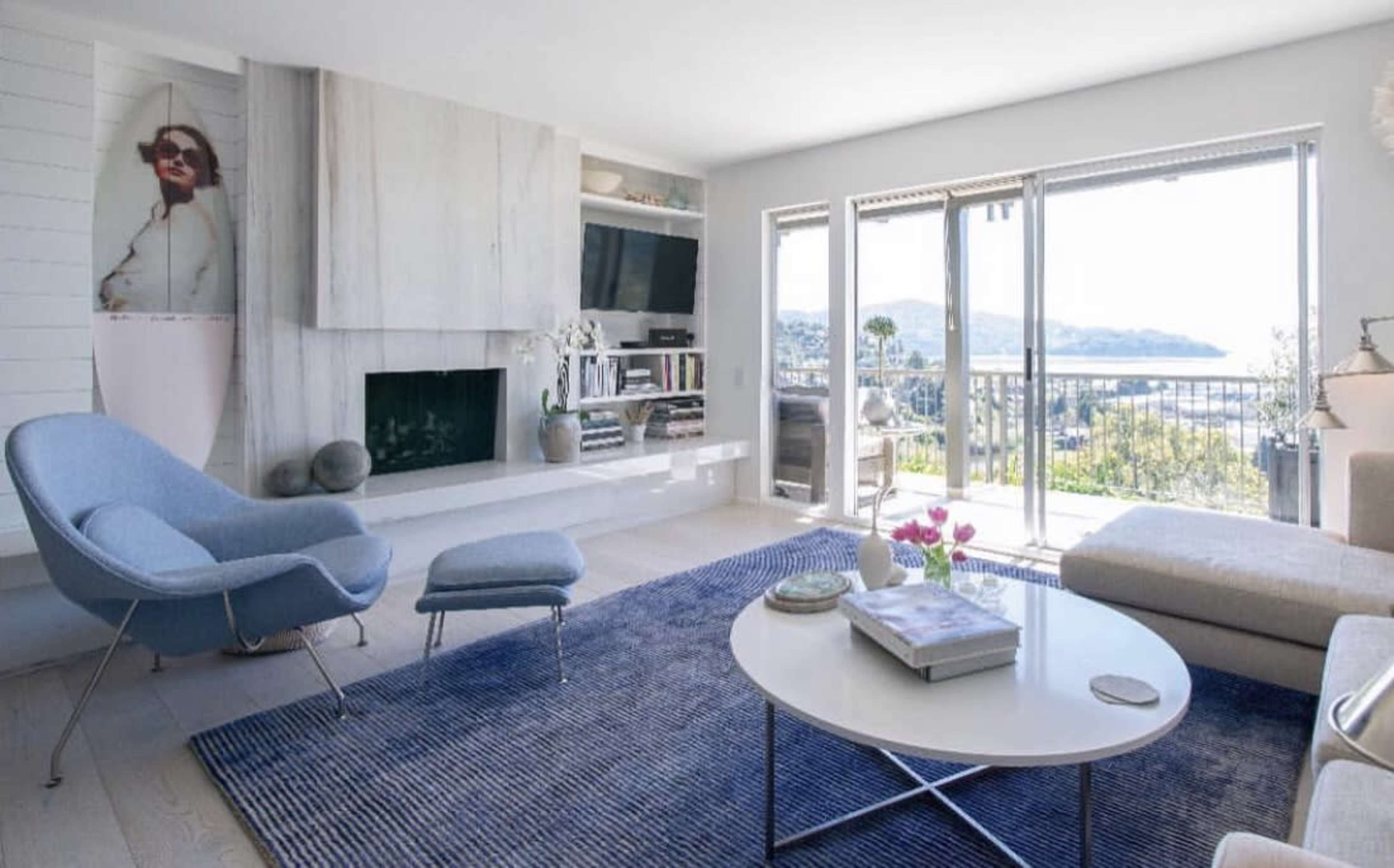 A modern living room with a large window overlooking a landscape, featuring a blue rug, a circular coffee table, a gray chair, and built-in shelves with various decorative items.