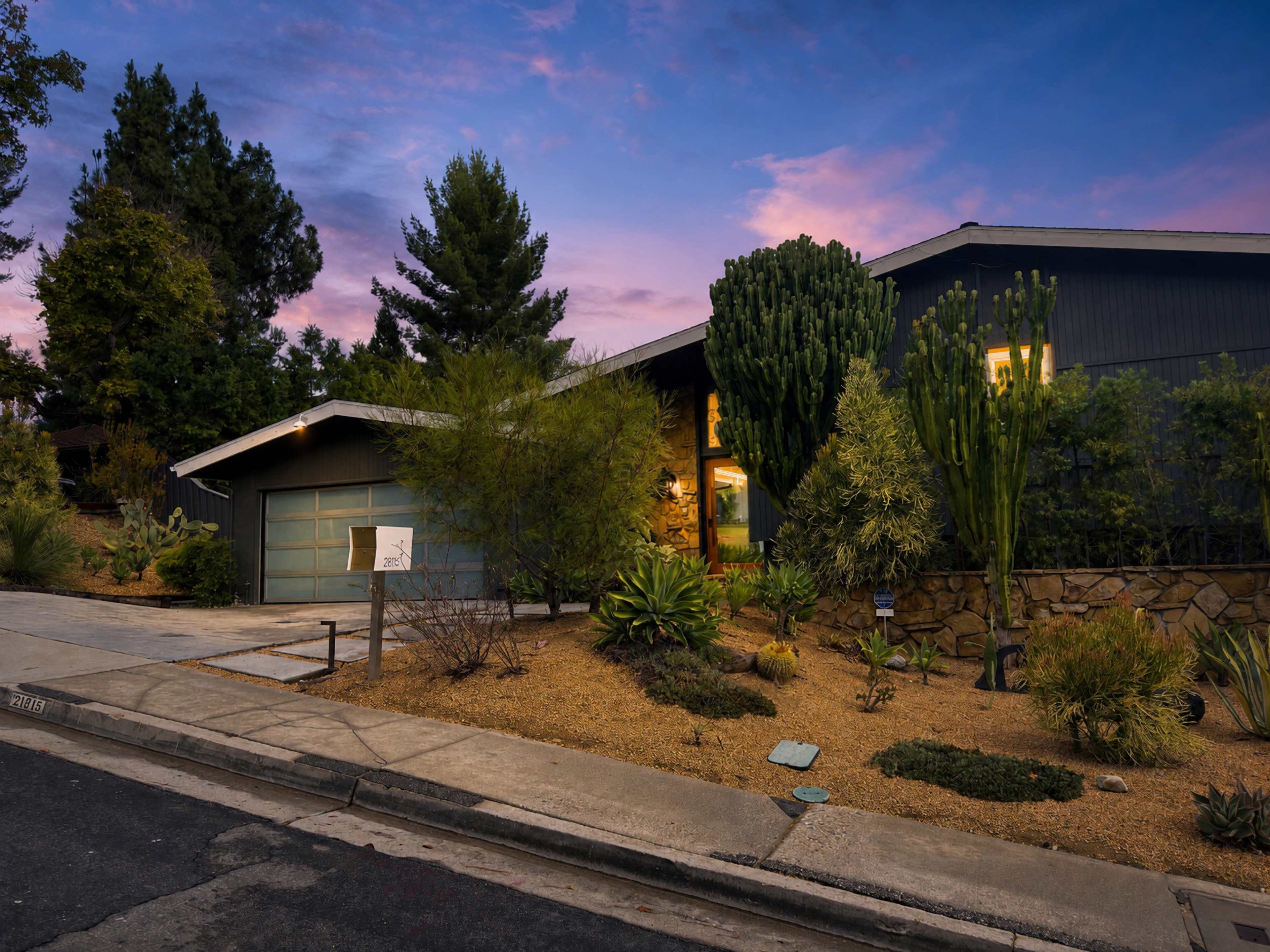 A modern house with a stone foundation and landscaped yard is situated at the end of a quiet street under a colorful sky.