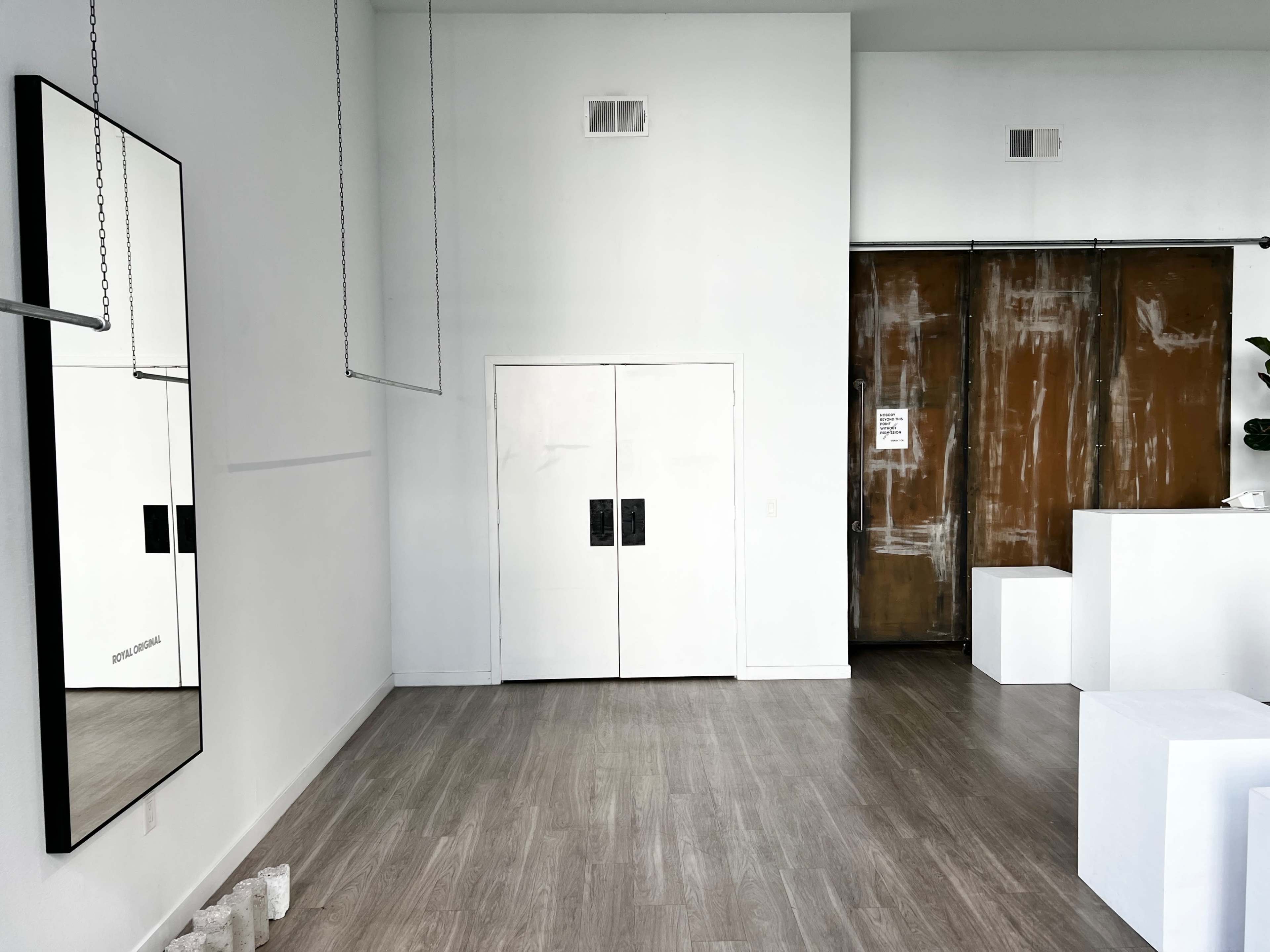 The image shows a minimalist interior space with light wood flooring, featuring a large mirror, two white double doors, and a rust-colored sliding door.
