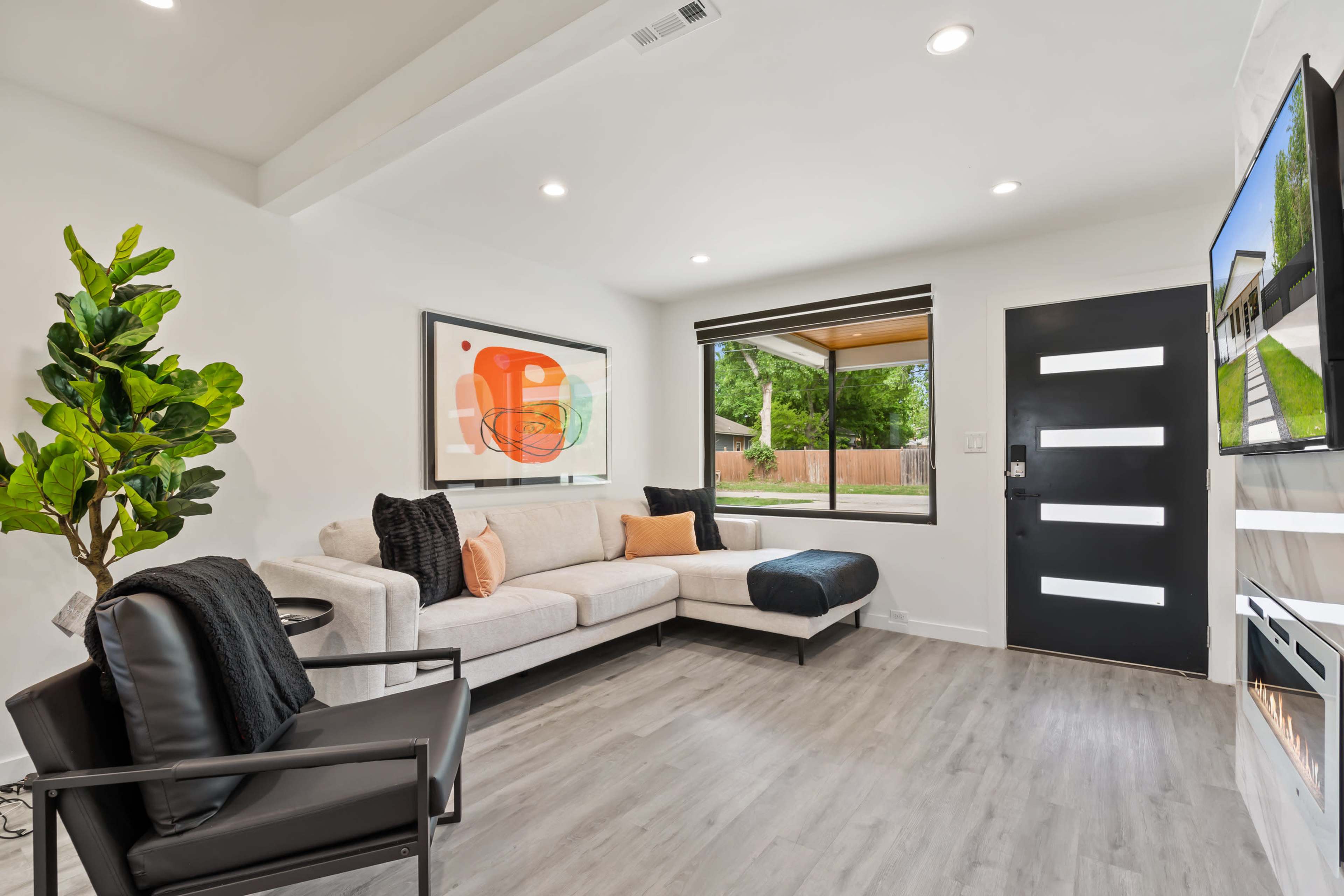 The modern living room features a light-colored sofa, a black armchair, a large window, and a digital fireplace wall.