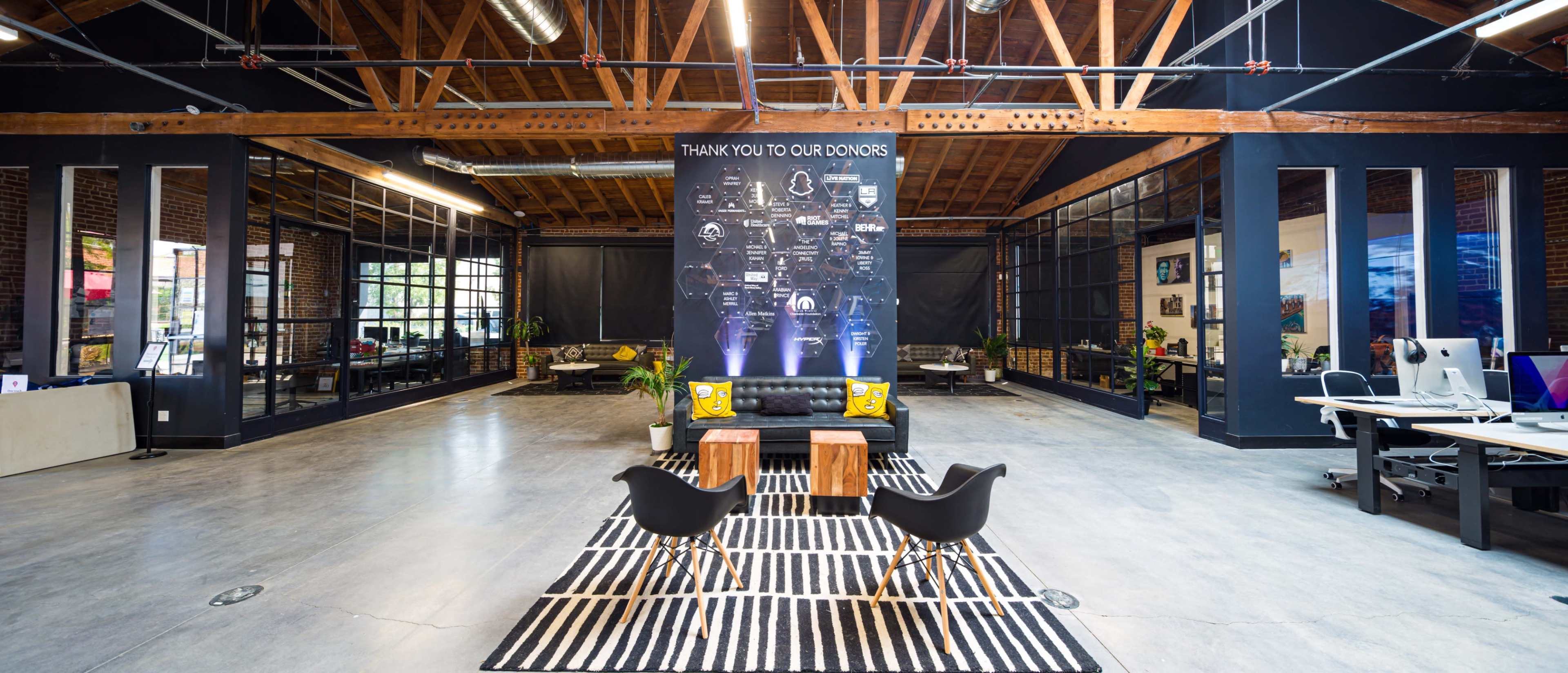A spacious modern office with exposed wooden beams, a seating area featuring black chairs and wooden tables, and a wall displaying acknowledgments to donors.