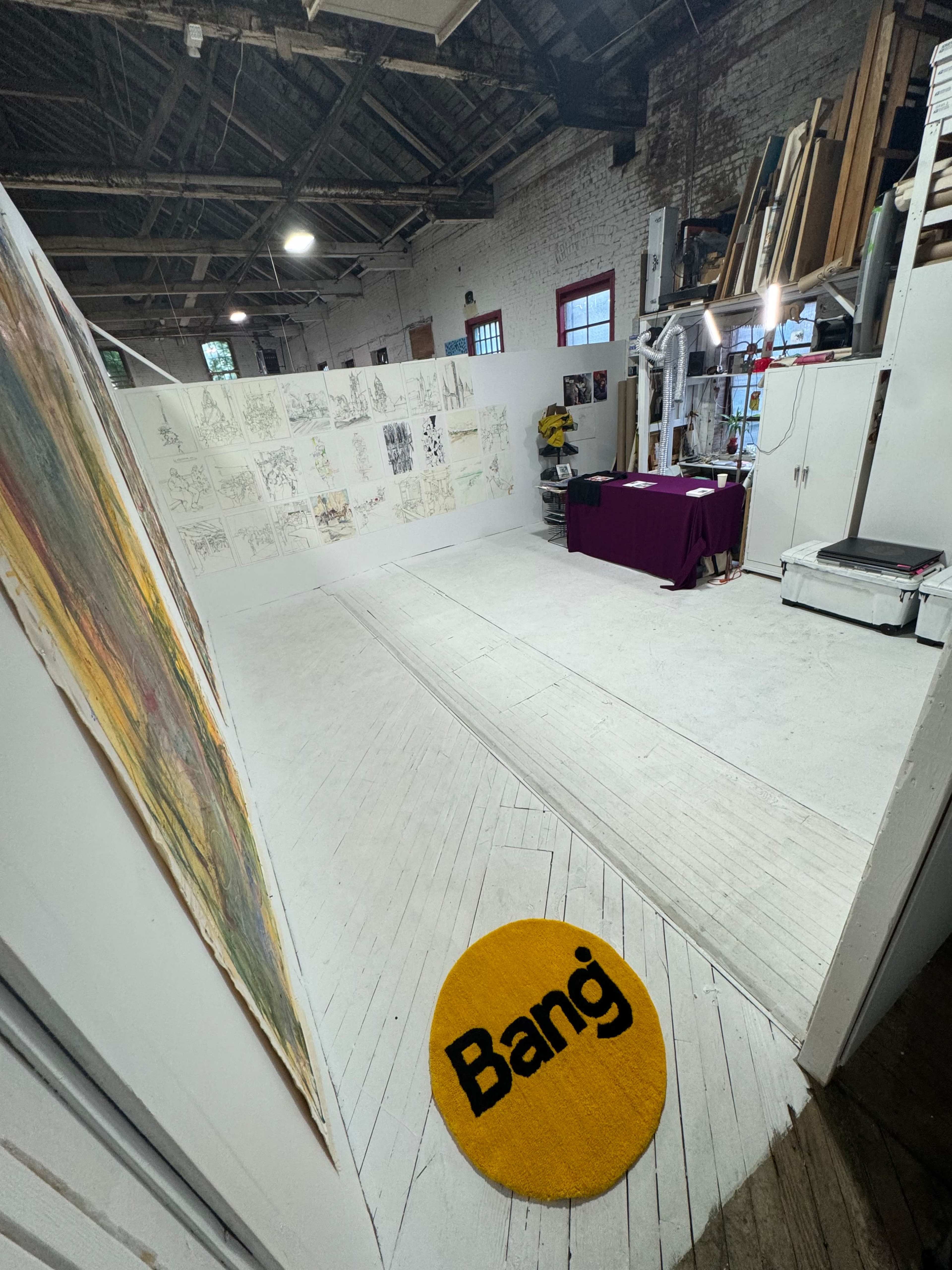 An art studio with white wooden floors, a large yellow rug with the word "Bang" in black, and walls covered with sketches and drawings.
