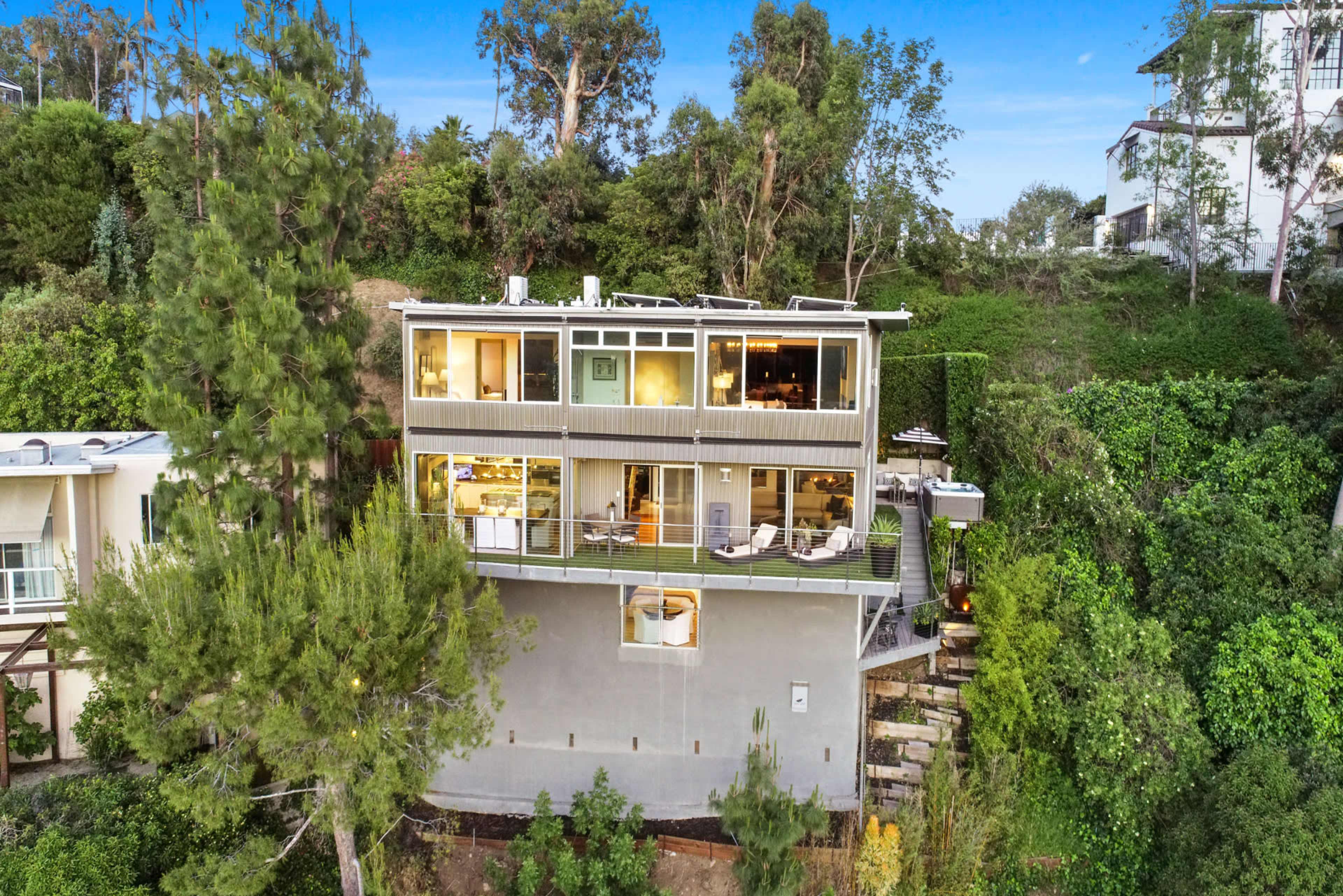 A modern multi-level house is situated on a hillside, surrounded by trees and greenery.