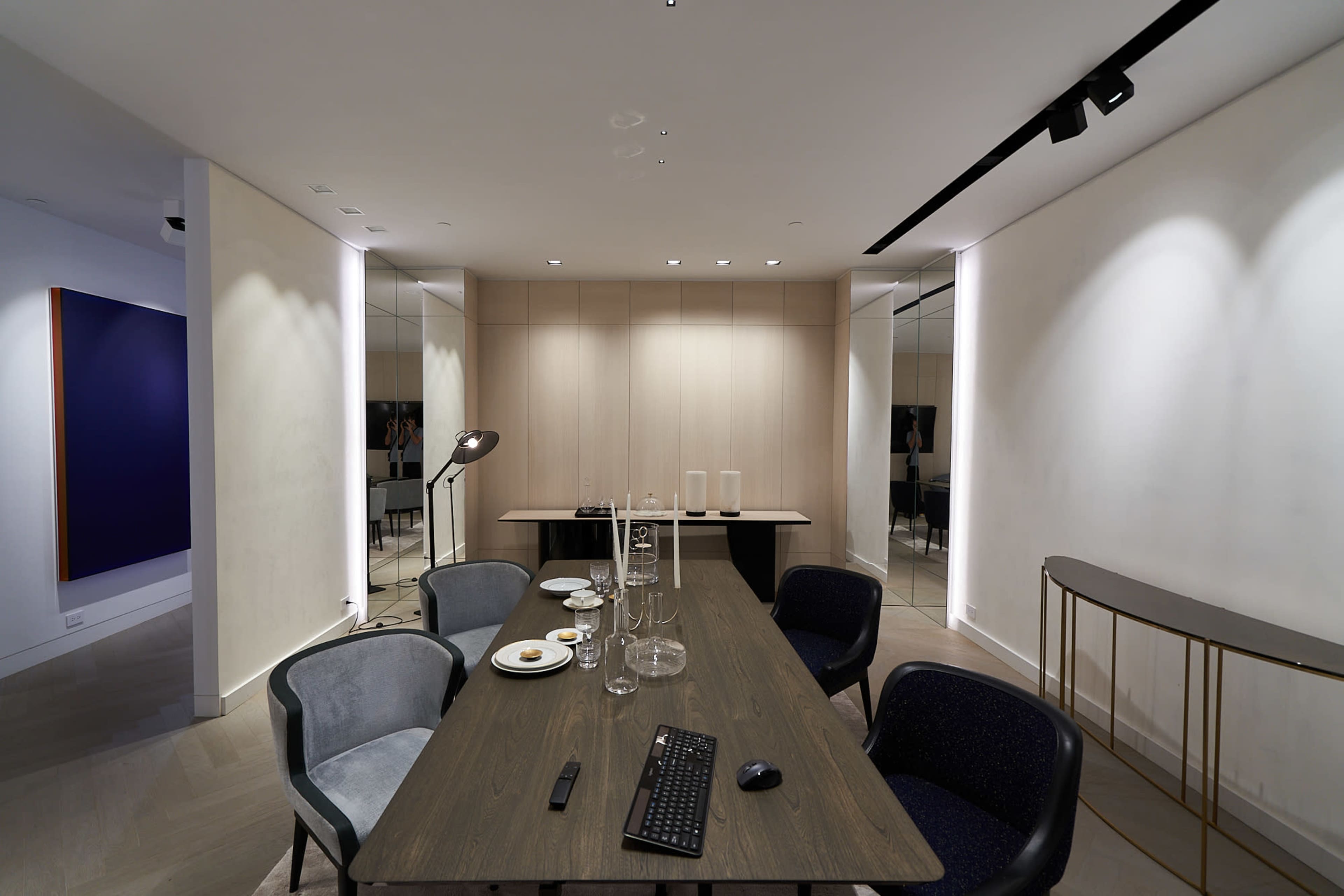 A conference room features a wooden table set with empty plates and glassware, surrounded by chairs, with mirrors and recessed lighting on the walls.