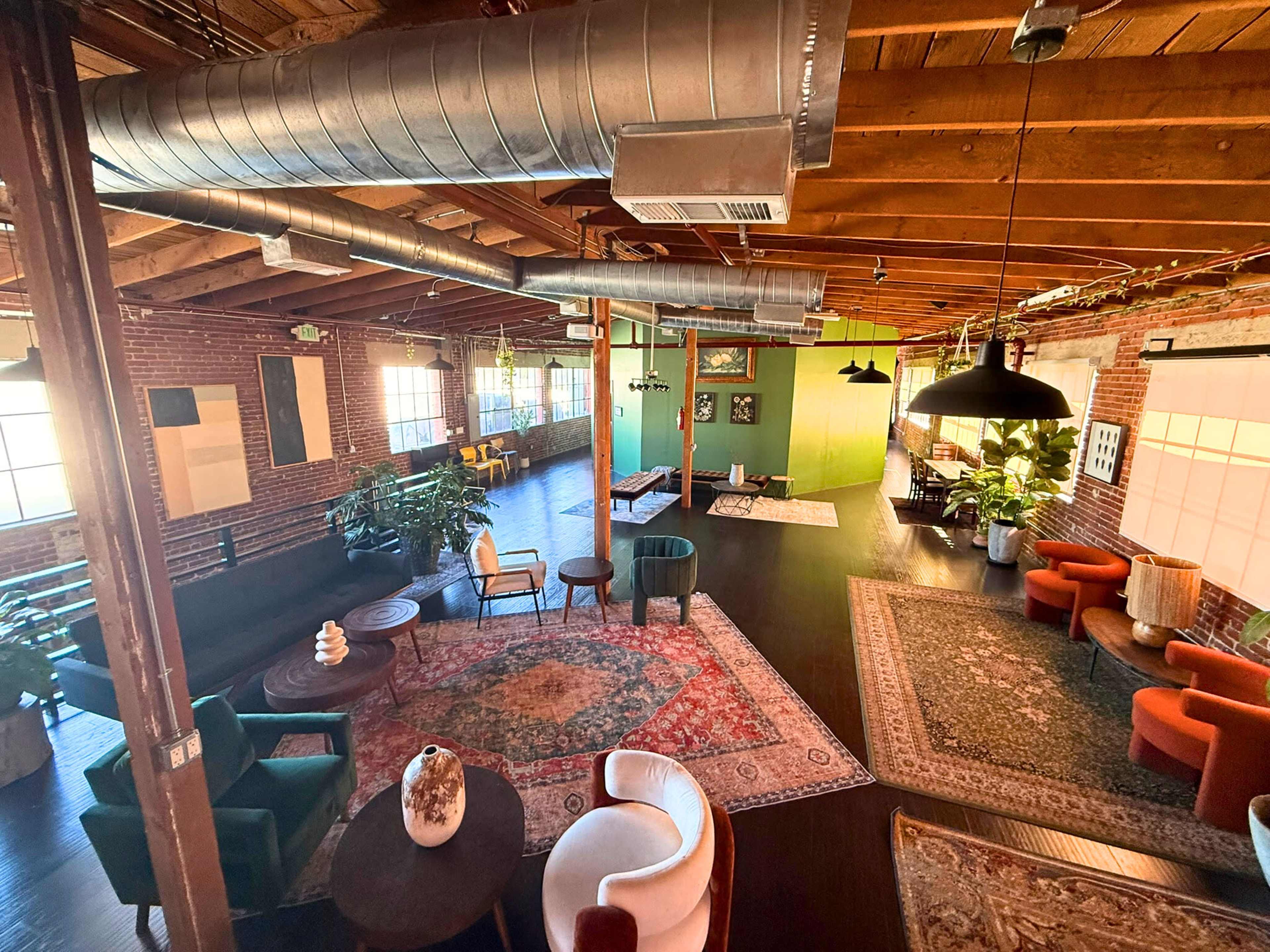 The image shows a spacious, modern interior with exposed wooden beams, large windows, various seating areas, and decorative rugs.