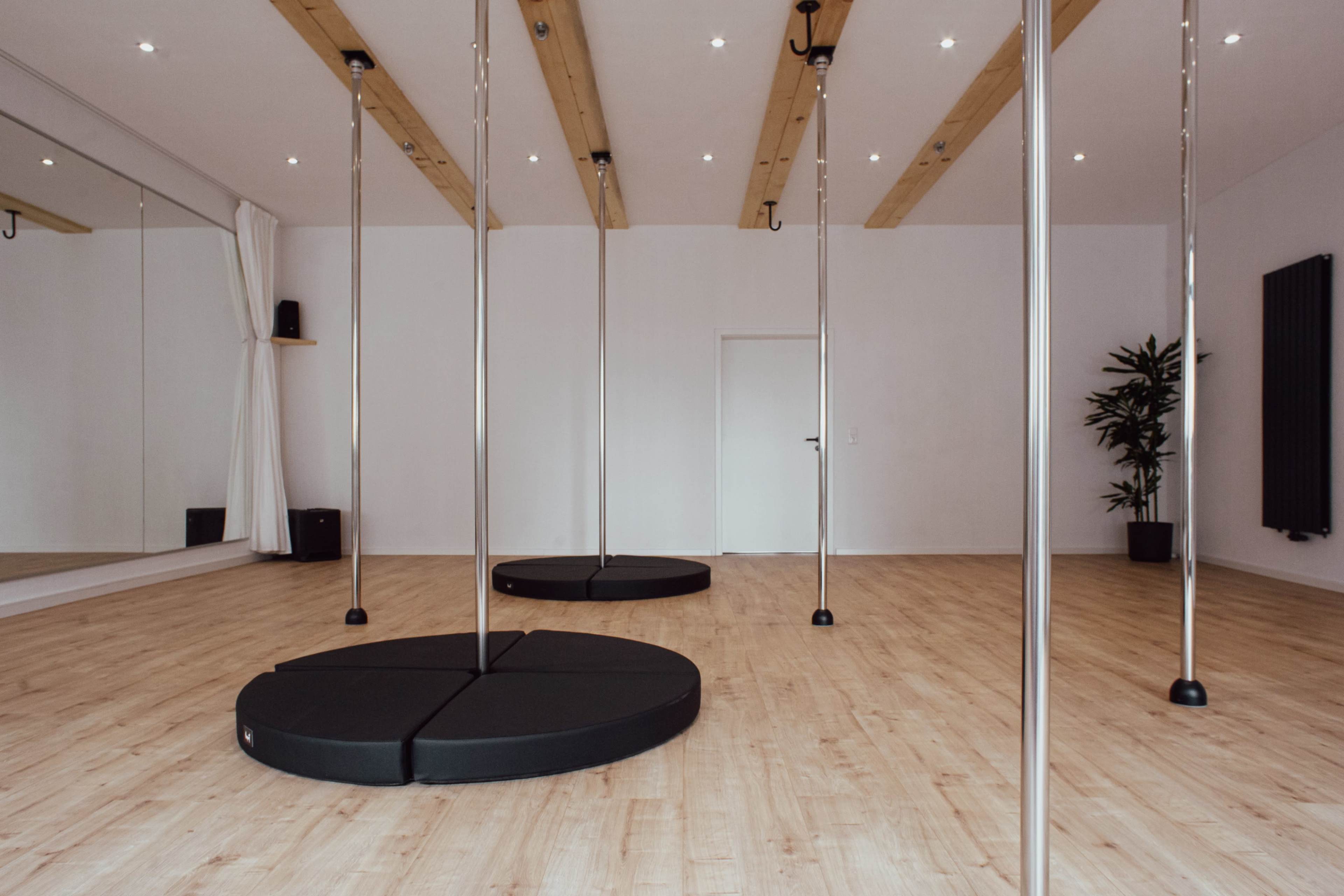 The image shows a spacious, brightly lit dance studio with three polished pole dance poles and a mirrored wall.