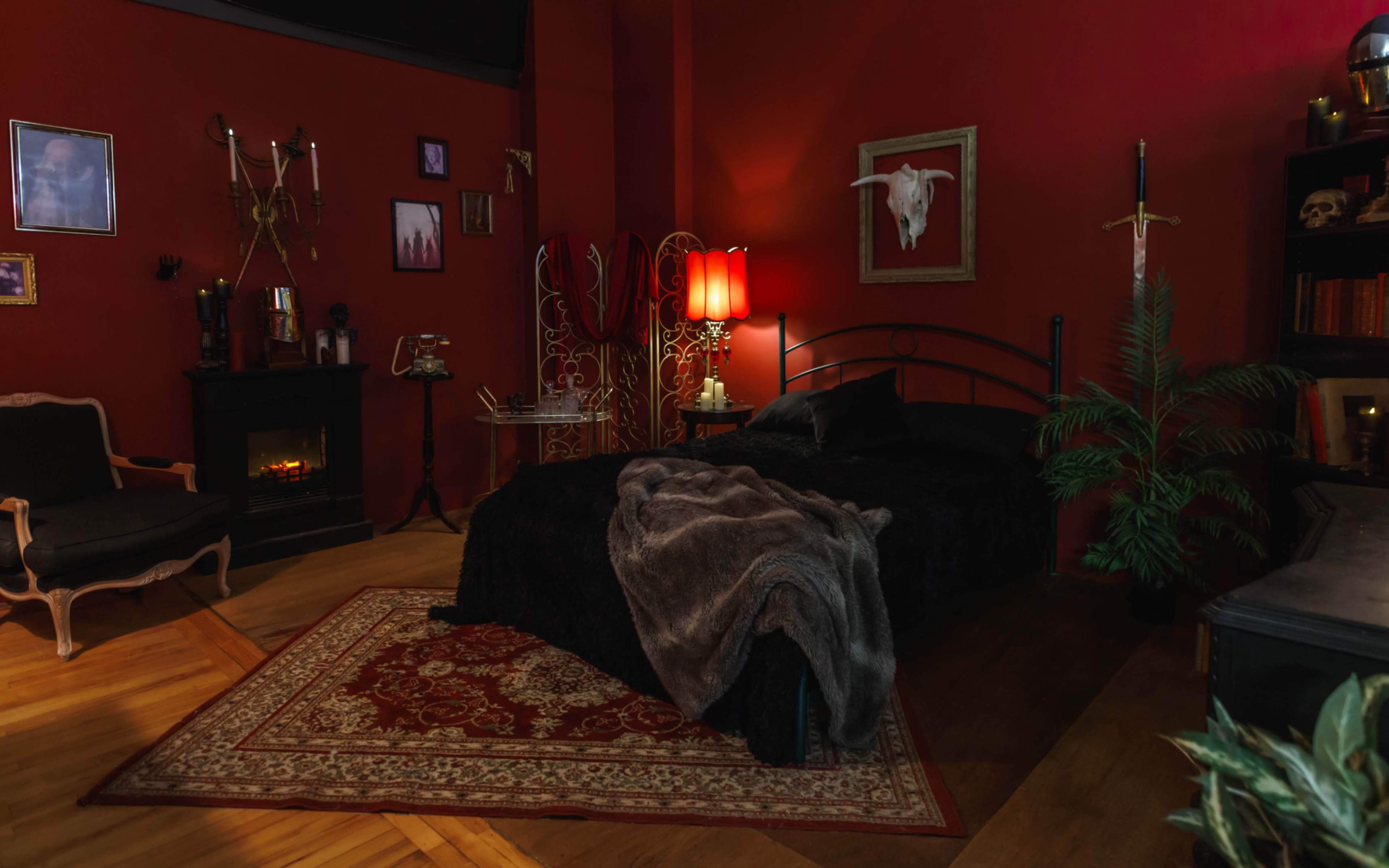 The scene depicts a dimly lit bedroom with red walls, a black bed covered in a fur blanket, a red lamp, and decorative elements, including a bull skull and assorted artwork.