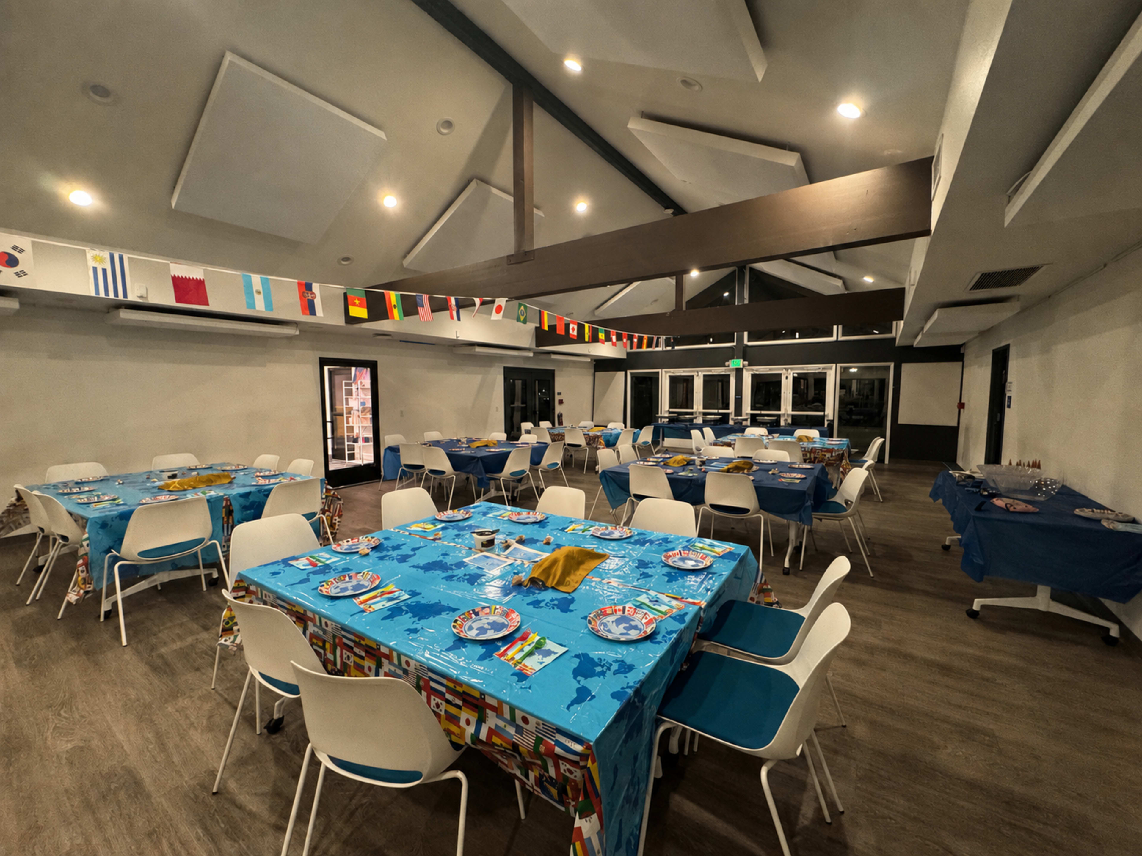 The image shows a dining area set up with multiple tables covered in blue tablecloths, decorated with plates and napkins, and featuring flags from various countries hanging from the ceiling.