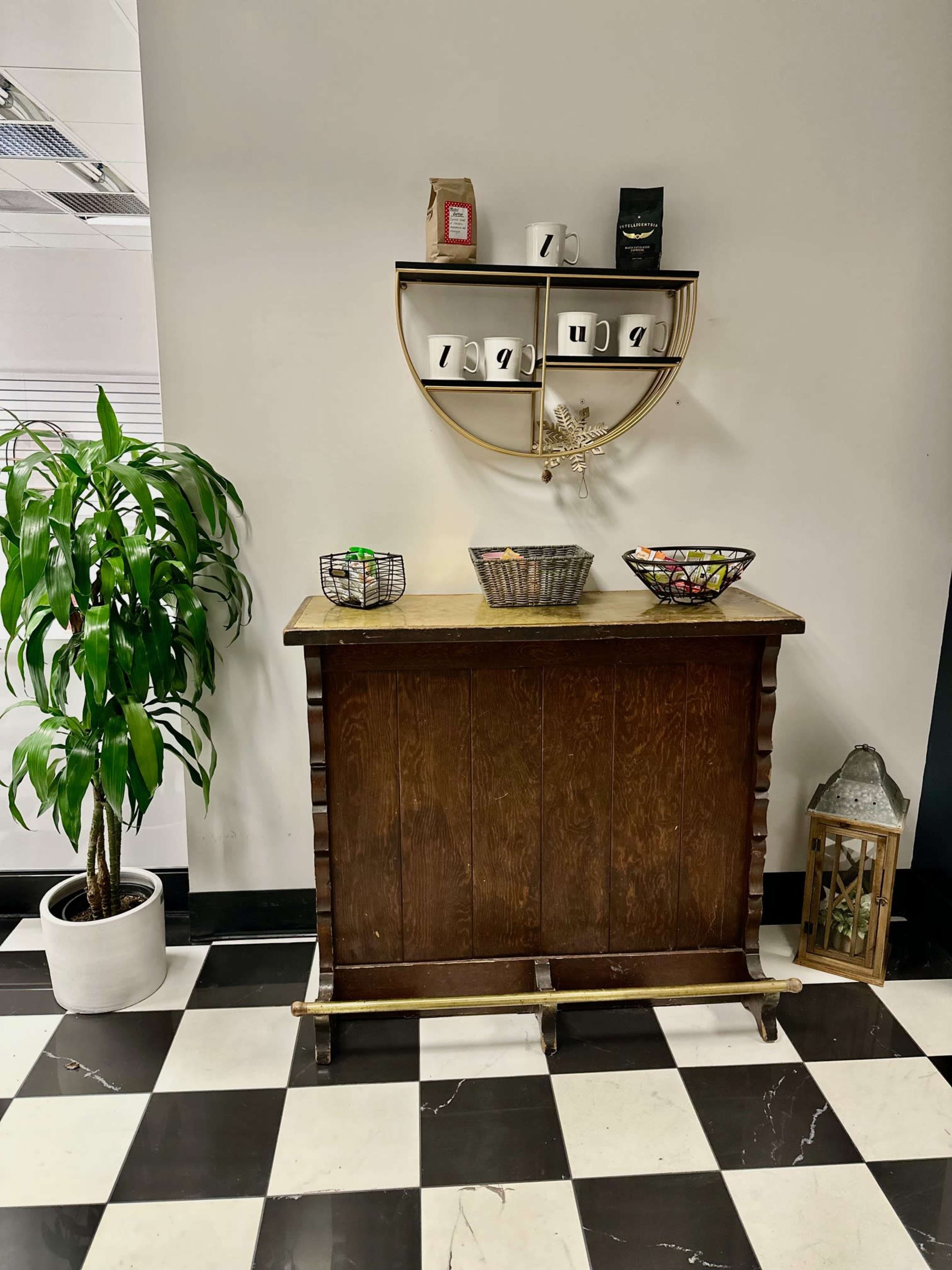 A wooden console table against a wall, with a plant beside it, decorative bowls on the table, and a shelf above displaying mugs and decorative items.