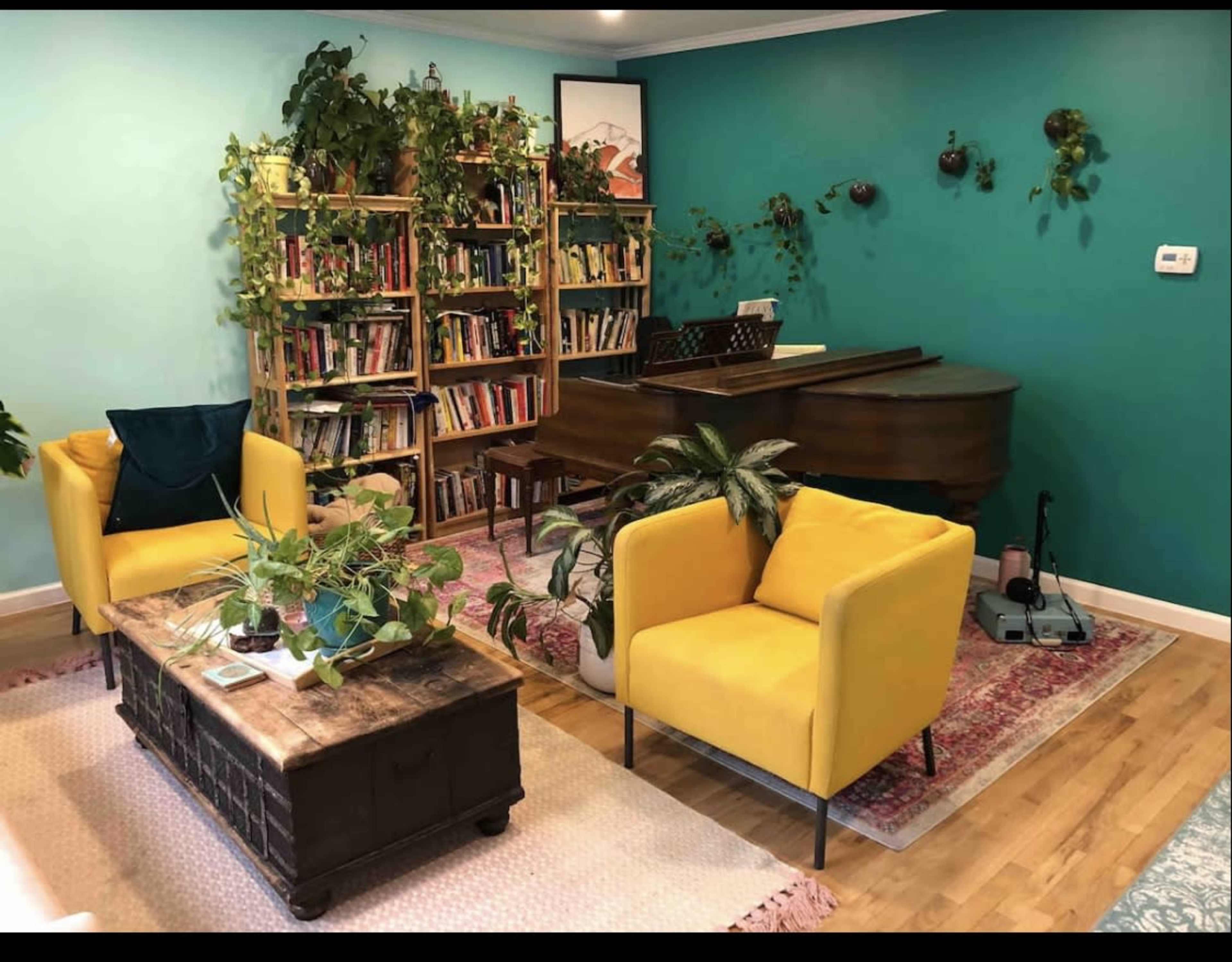 A bright living room features a piano, a bookshelf filled with books, two yellow chairs, and various potted plants against a teal wall.