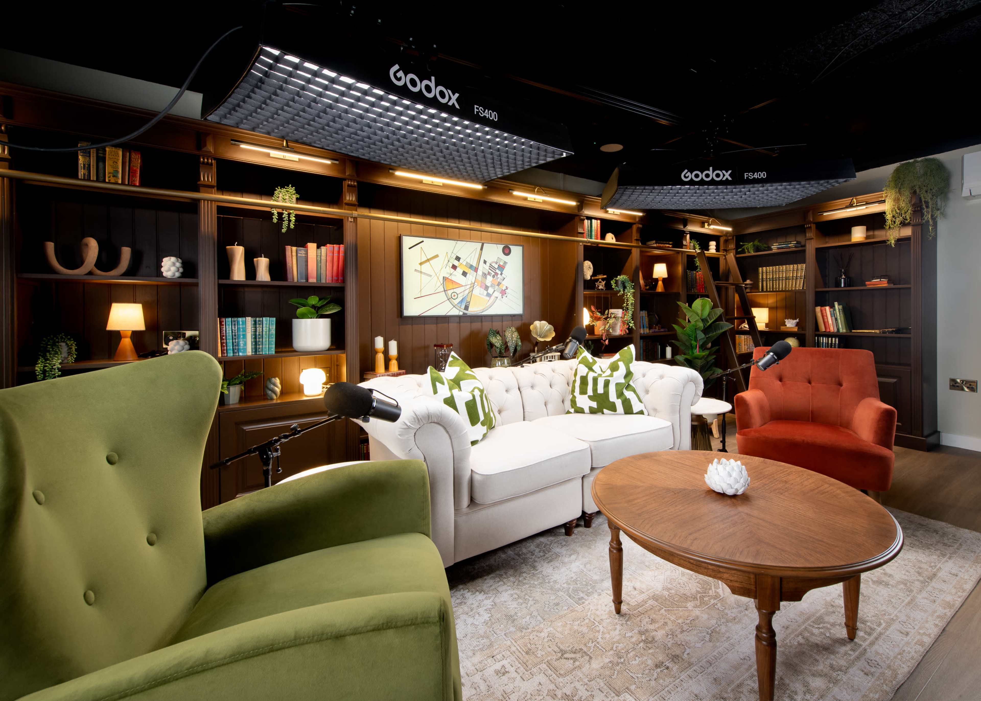 The image features a modern interview space with two distinct chairs, a white sofa, a round wooden coffee table, and a bookshelf filled with decorative items and plants.
