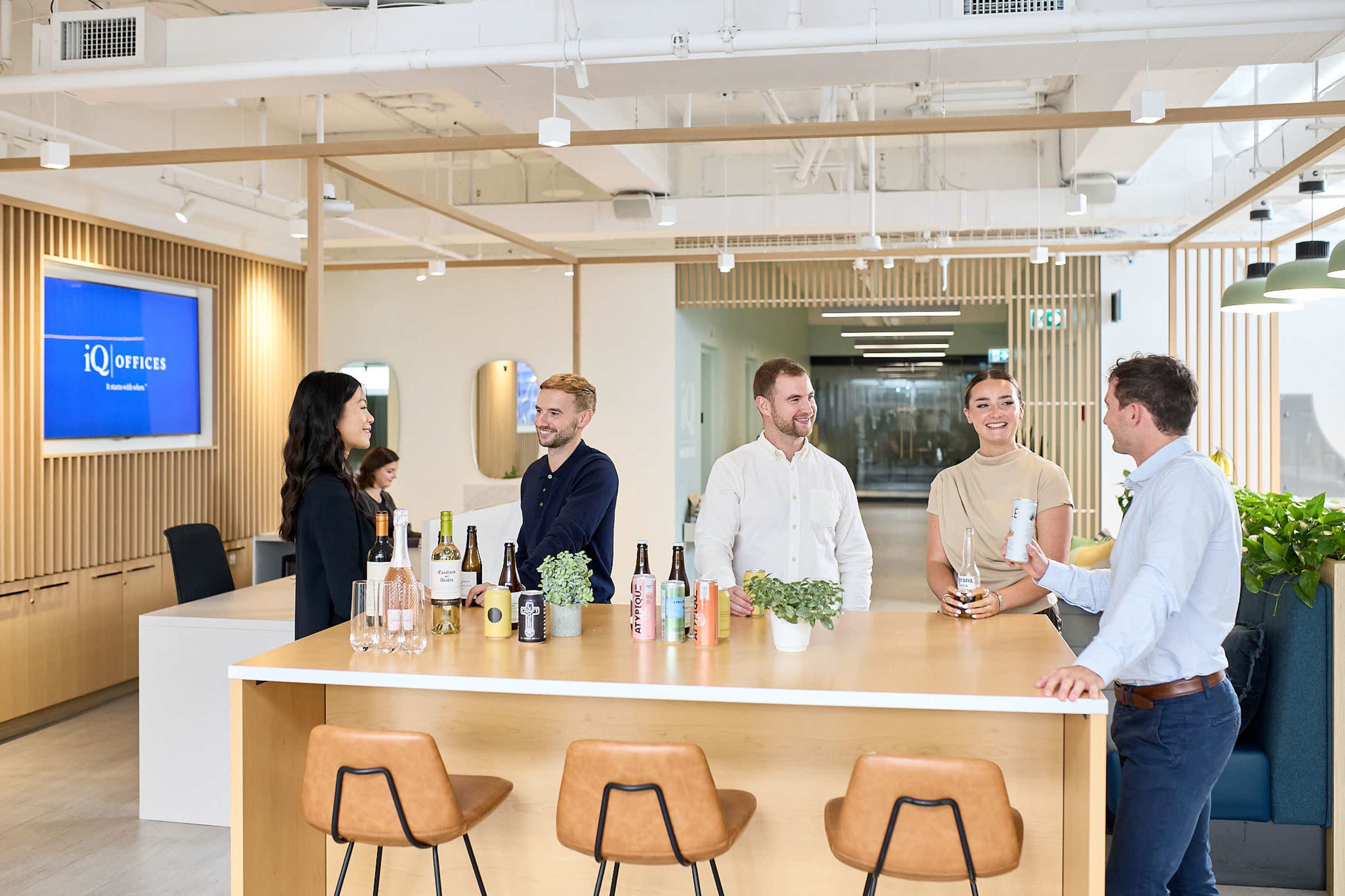 A group of four people chats and shares drinks at a modern office lounge with a light, open design.