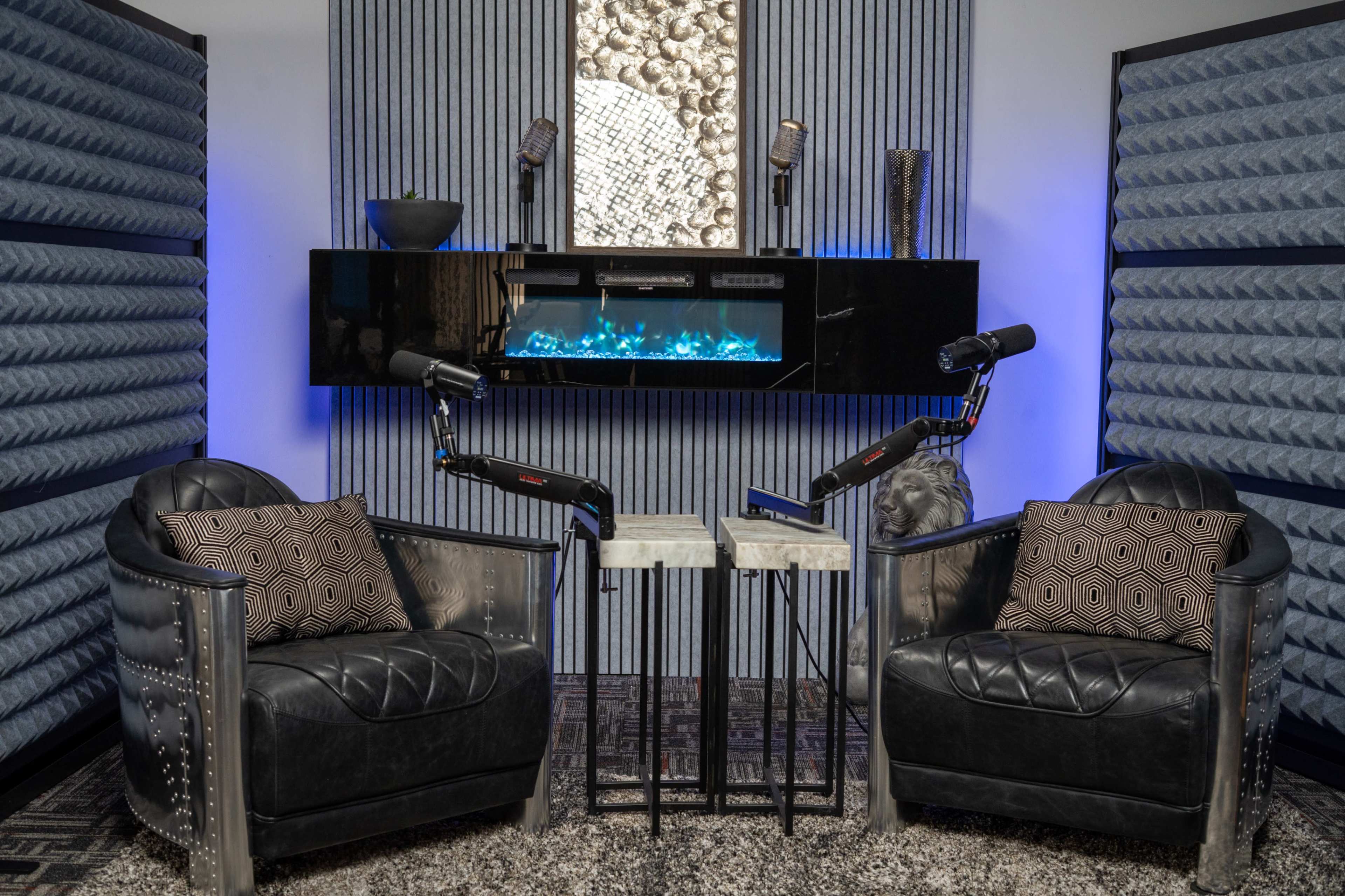 The image features a modern podcast studio with two black leather chairs, a small table between them, and wall-mounted microphones, all set against soundproofed panels and a decorative lighted aquarium.