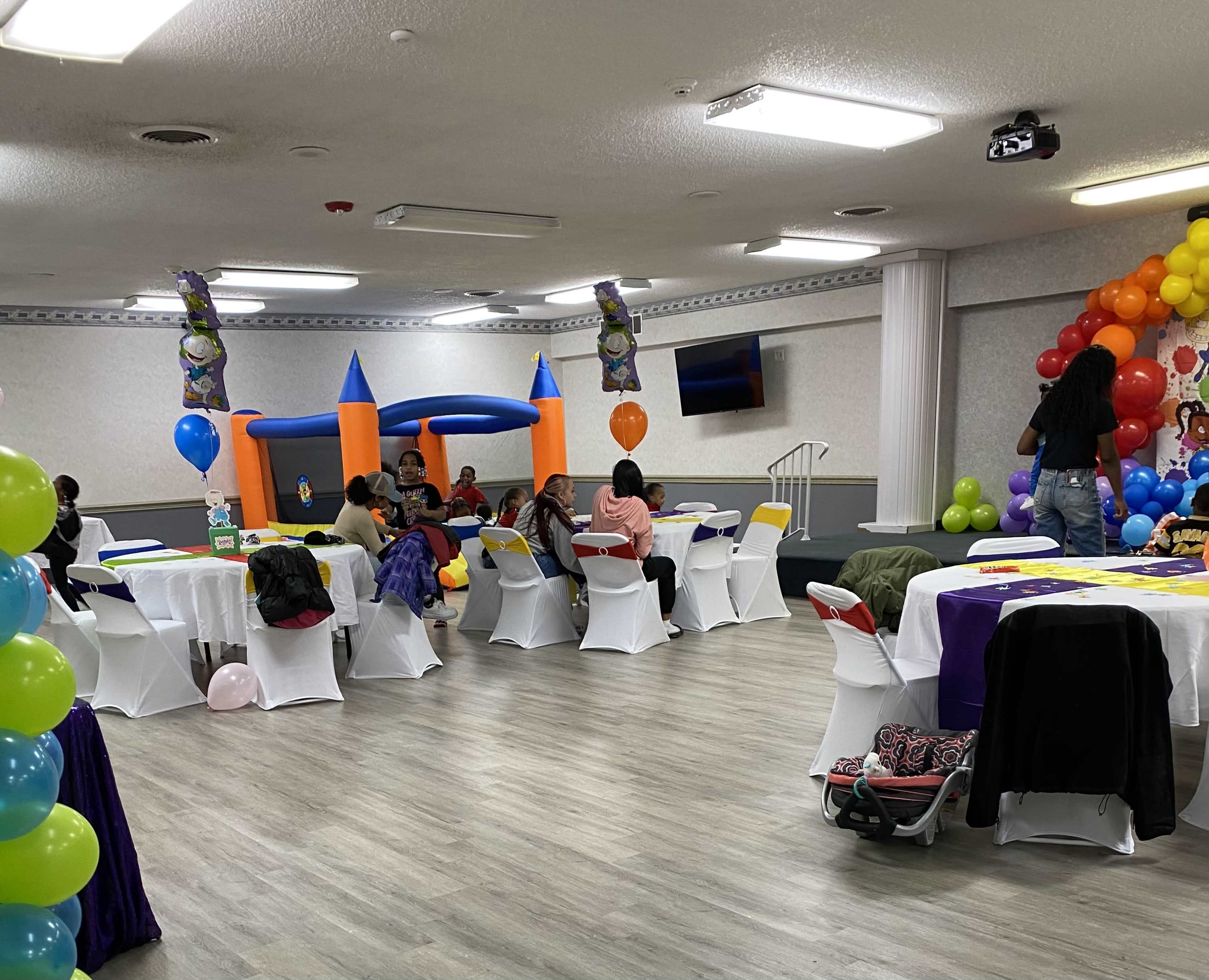A party or event space with several round tables covered in white tablecloths, colorful decorations, a bounce house in the background, and guests mingling throughout the room.