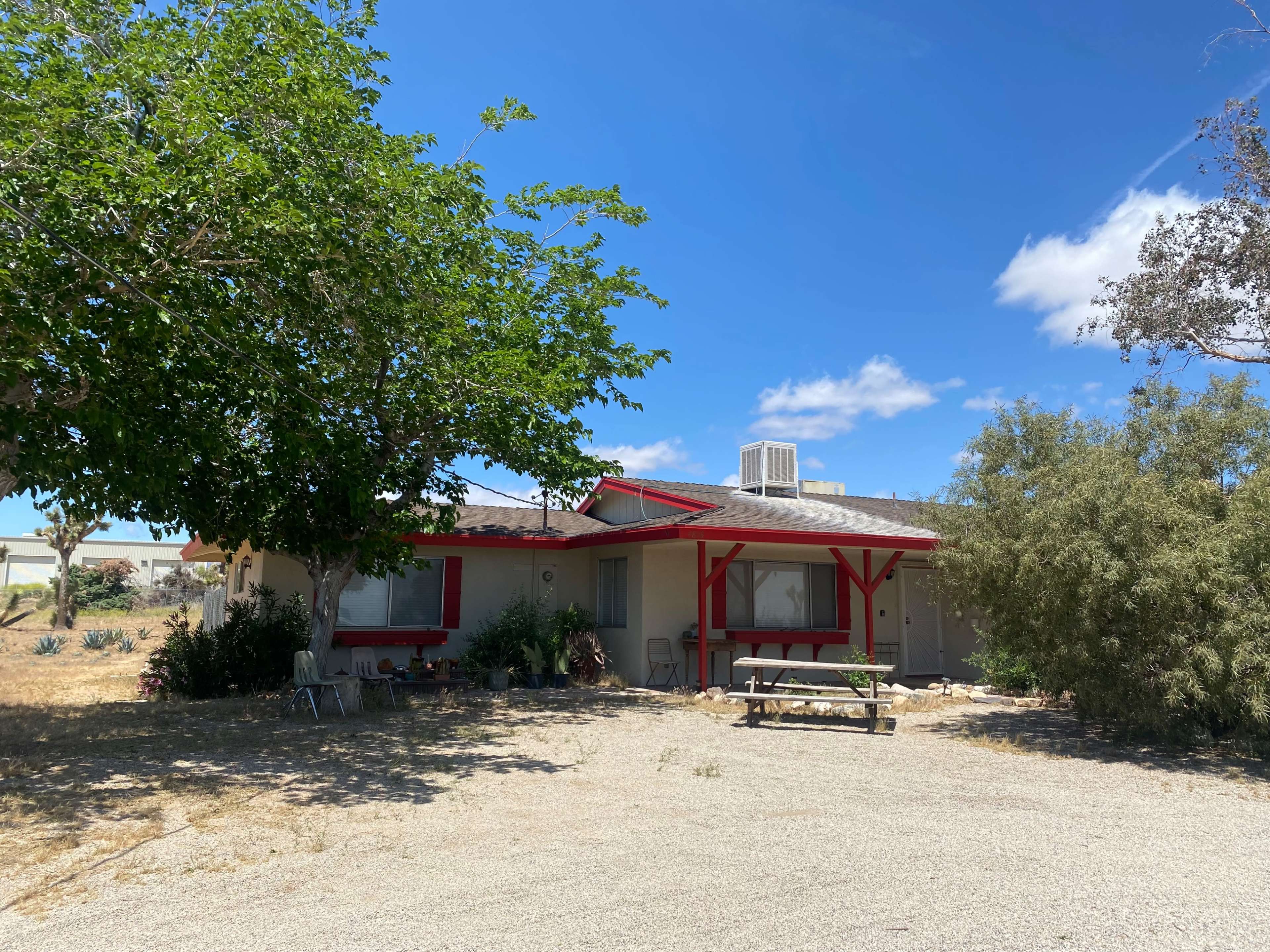 A single-story house with red trim is surrounded by gravel and a few trees in a semi-arid landscape.