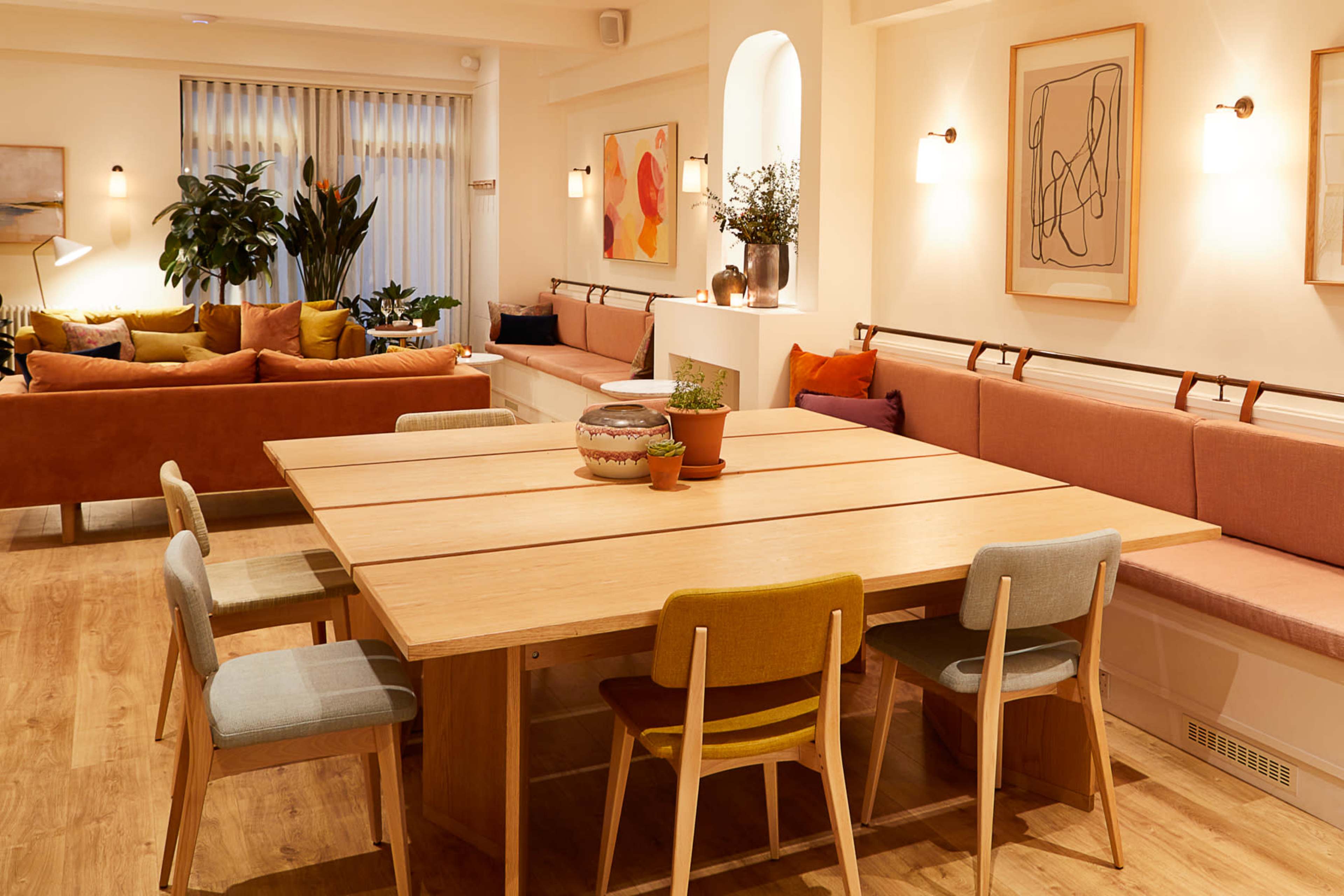 A cozy interior space featuring a large wooden table surrounded by colorful chairs, with a bench along the wall and various plants and artwork decoratively placed.