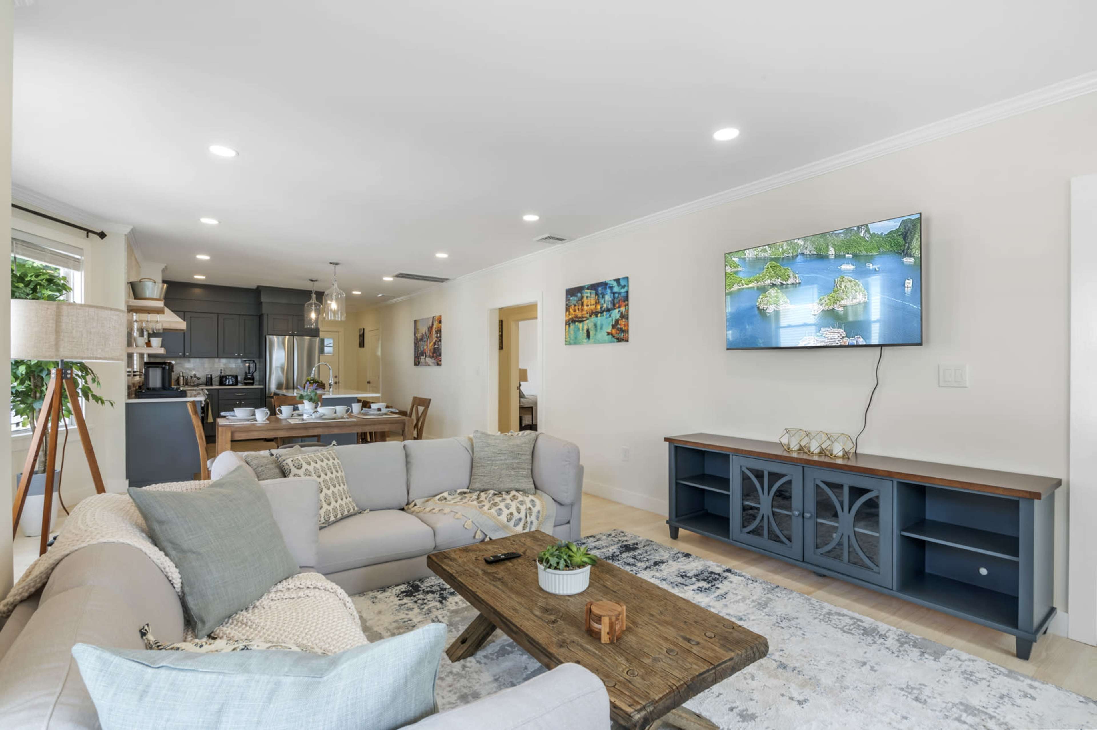 The living room features a light-colored sofa, a wooden coffee table, and a wall-mounted television, with a dining area visible in the background.