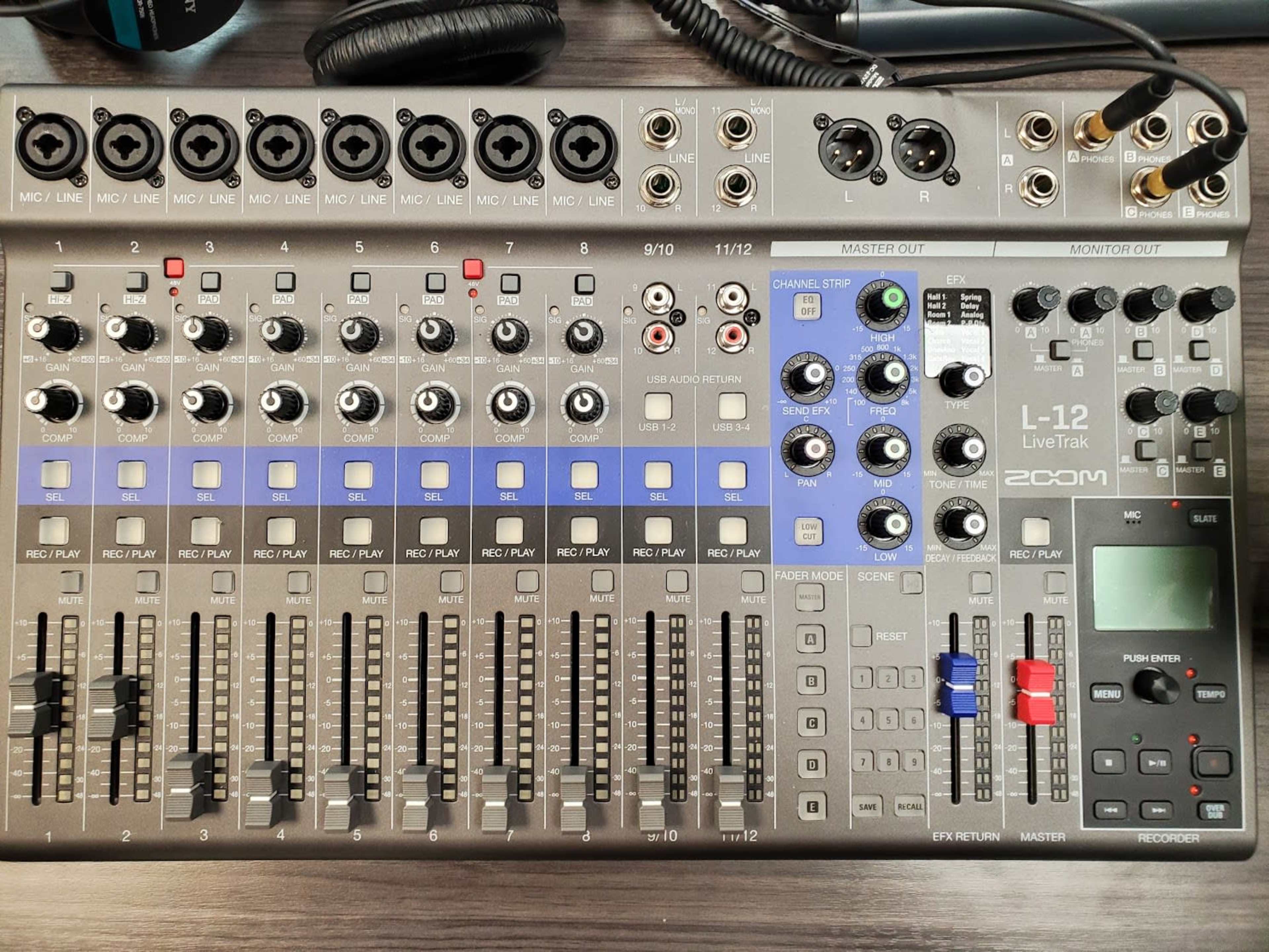 A Zoom L-12 LiveTrak audio mixing console with various channels, knobs, and output ports arranged on a desk.