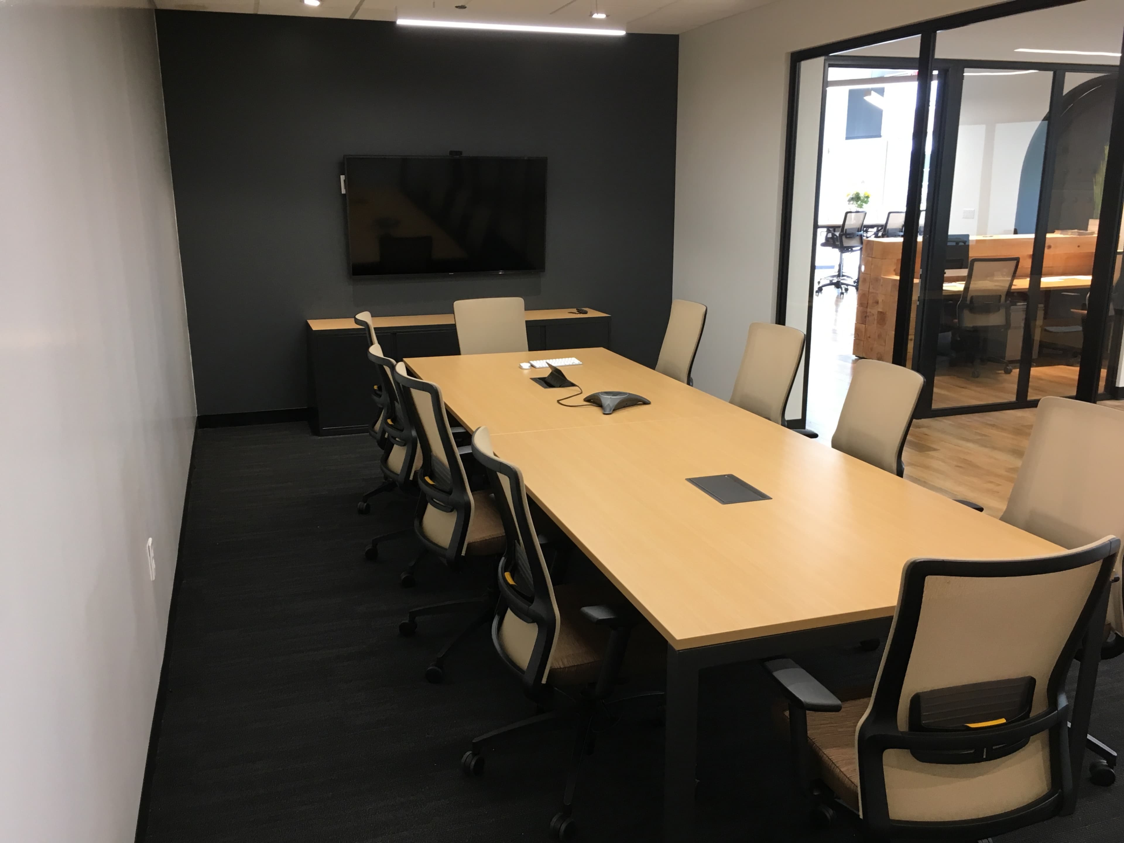 A modern conference room features a long wooden table surrounded by ergonomic chairs, with a large screen mounted on the wall and glass walls separating it from adjacent offices.