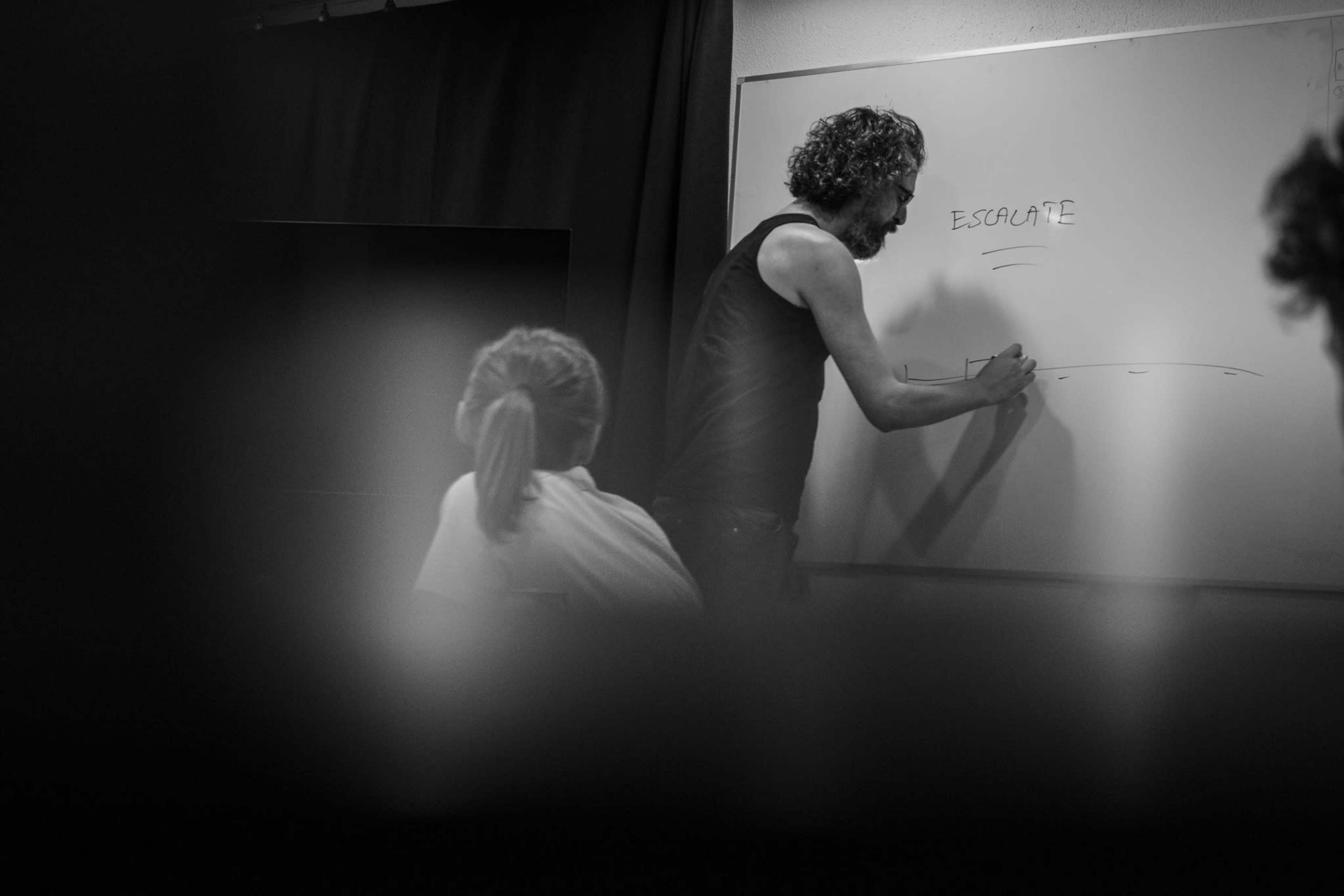 A man is standing at a whiteboard, writing the word "ESCALATE," while a person seated in the foreground observes him.