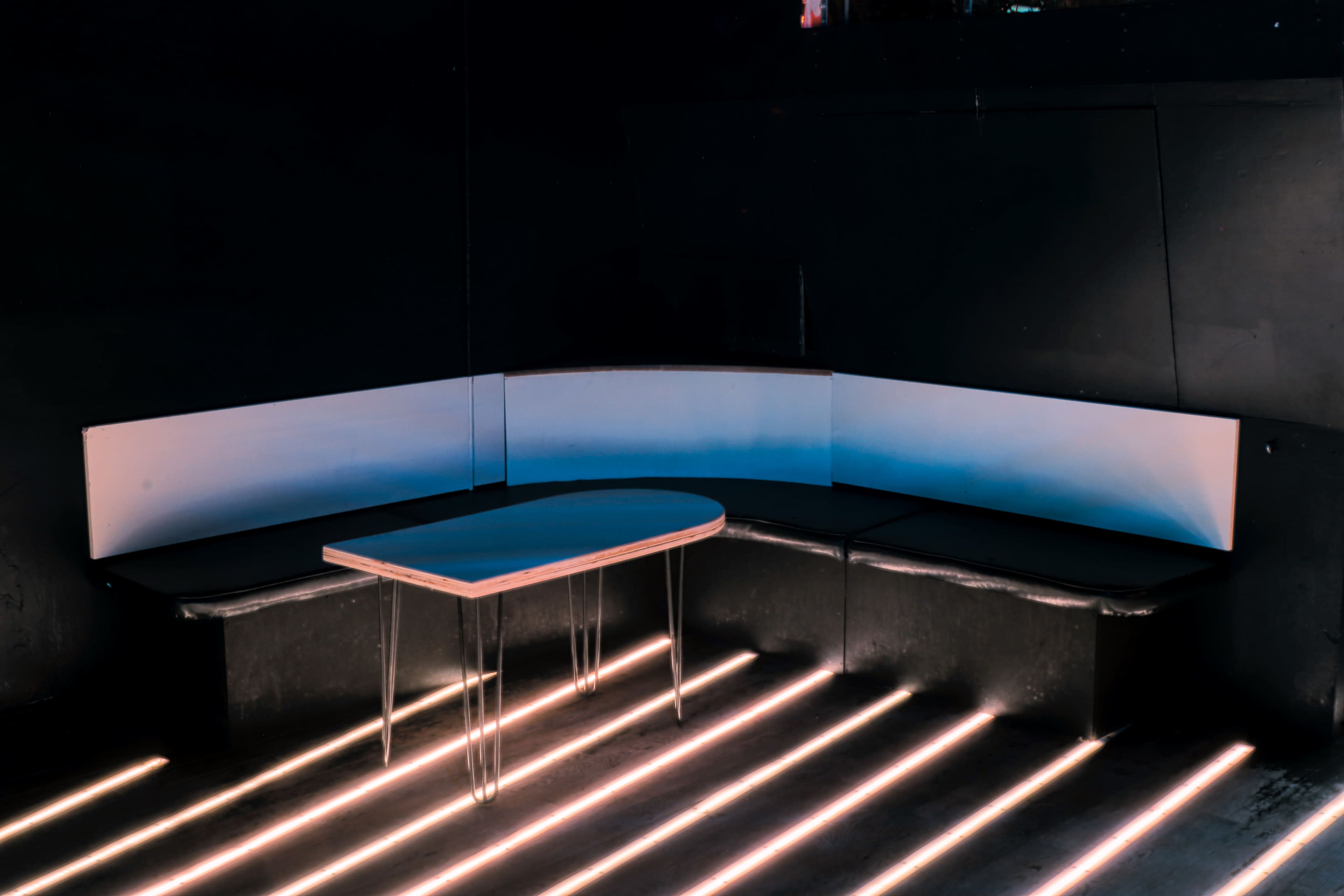 A dimly lit corner of a room features a curved seating area with a wooden table and illuminated floors.
