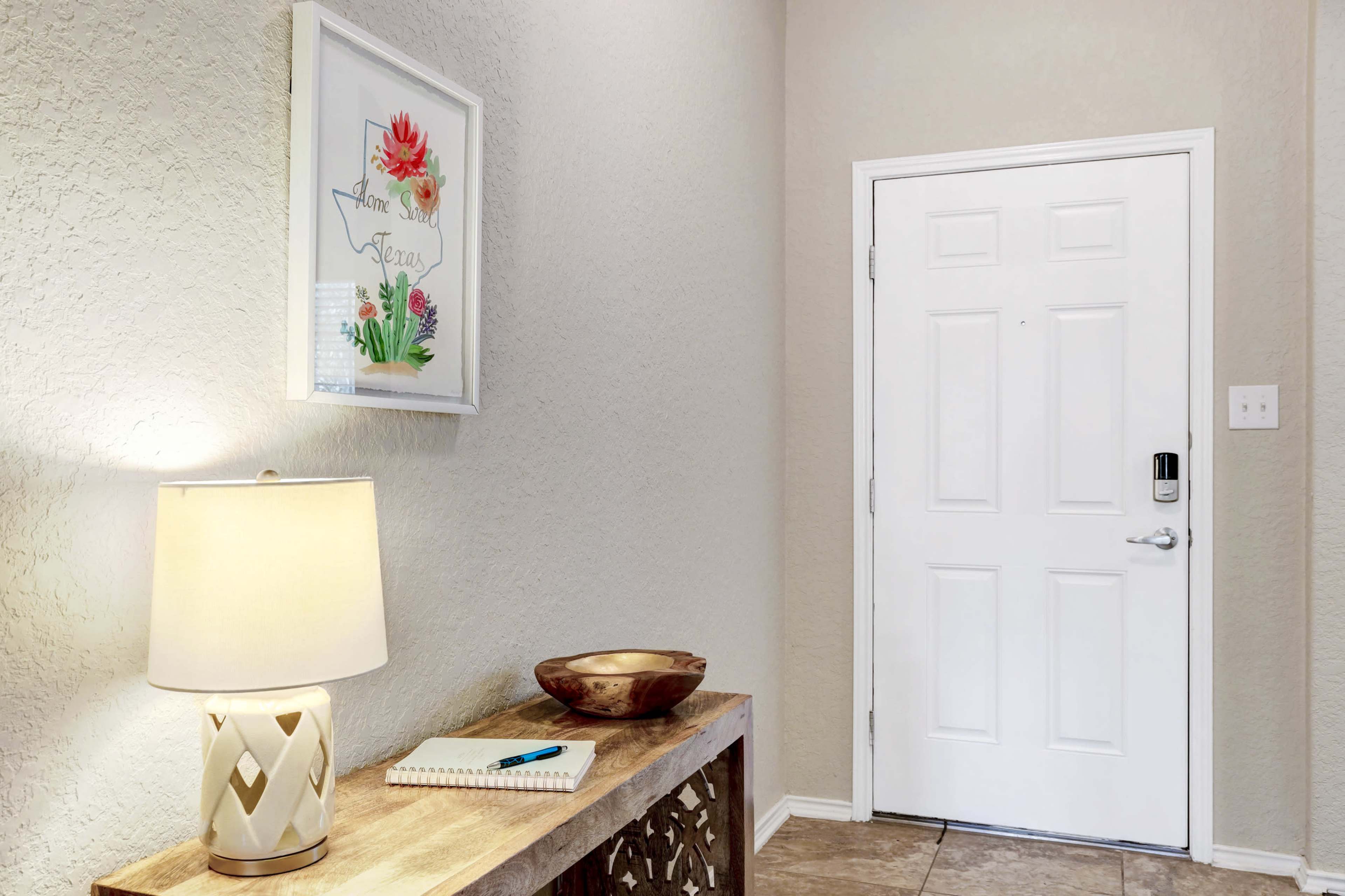 A light-colored hallway features a decorative console table with a lamp, a bowl, and a notebook, alongside a framed floral print on the wall and a white door.
