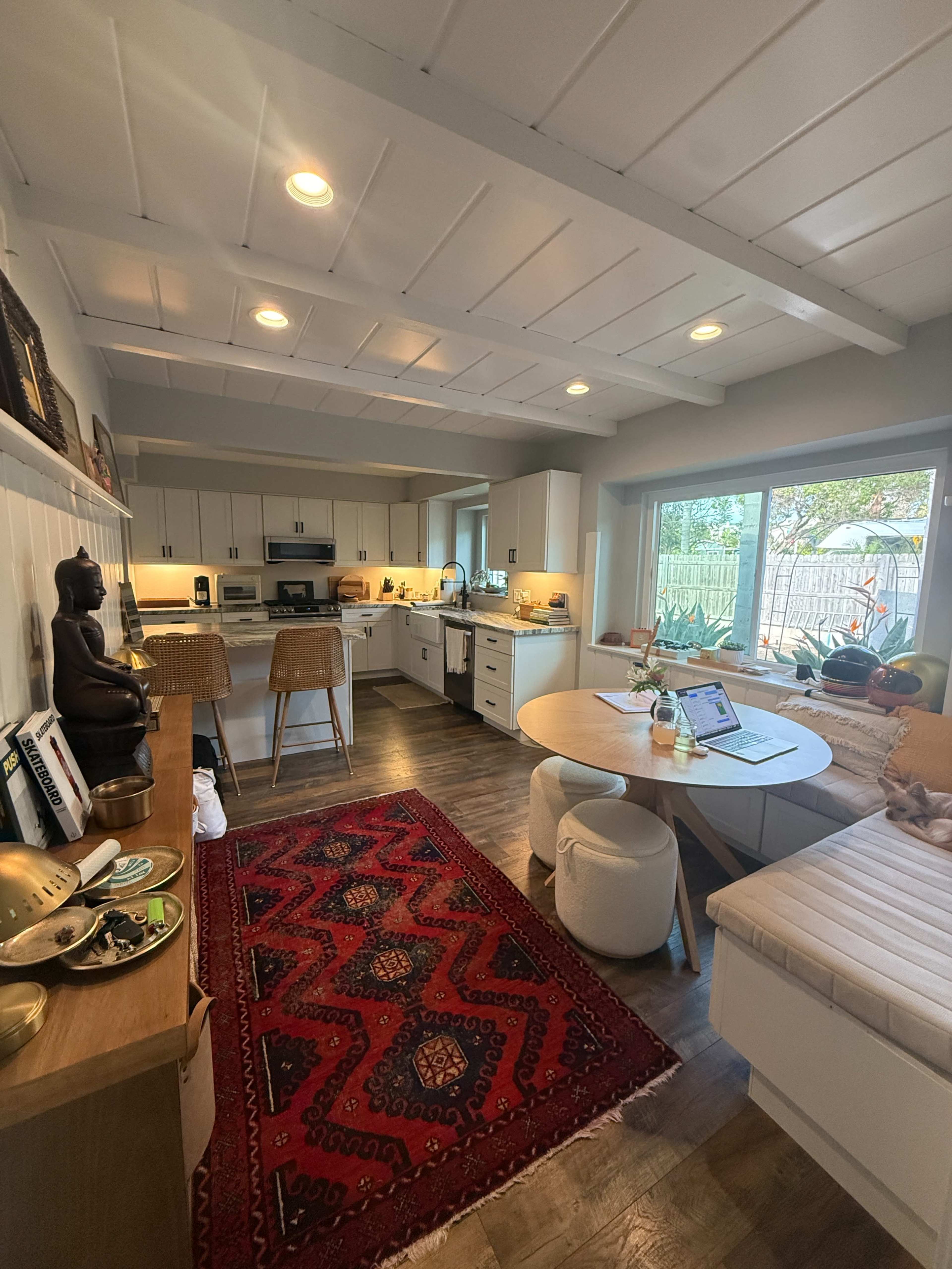 A bright kitchen features white cabinetry, a round dining table, and a mix of modern and traditional decor, including a large window and a patterned rug.
