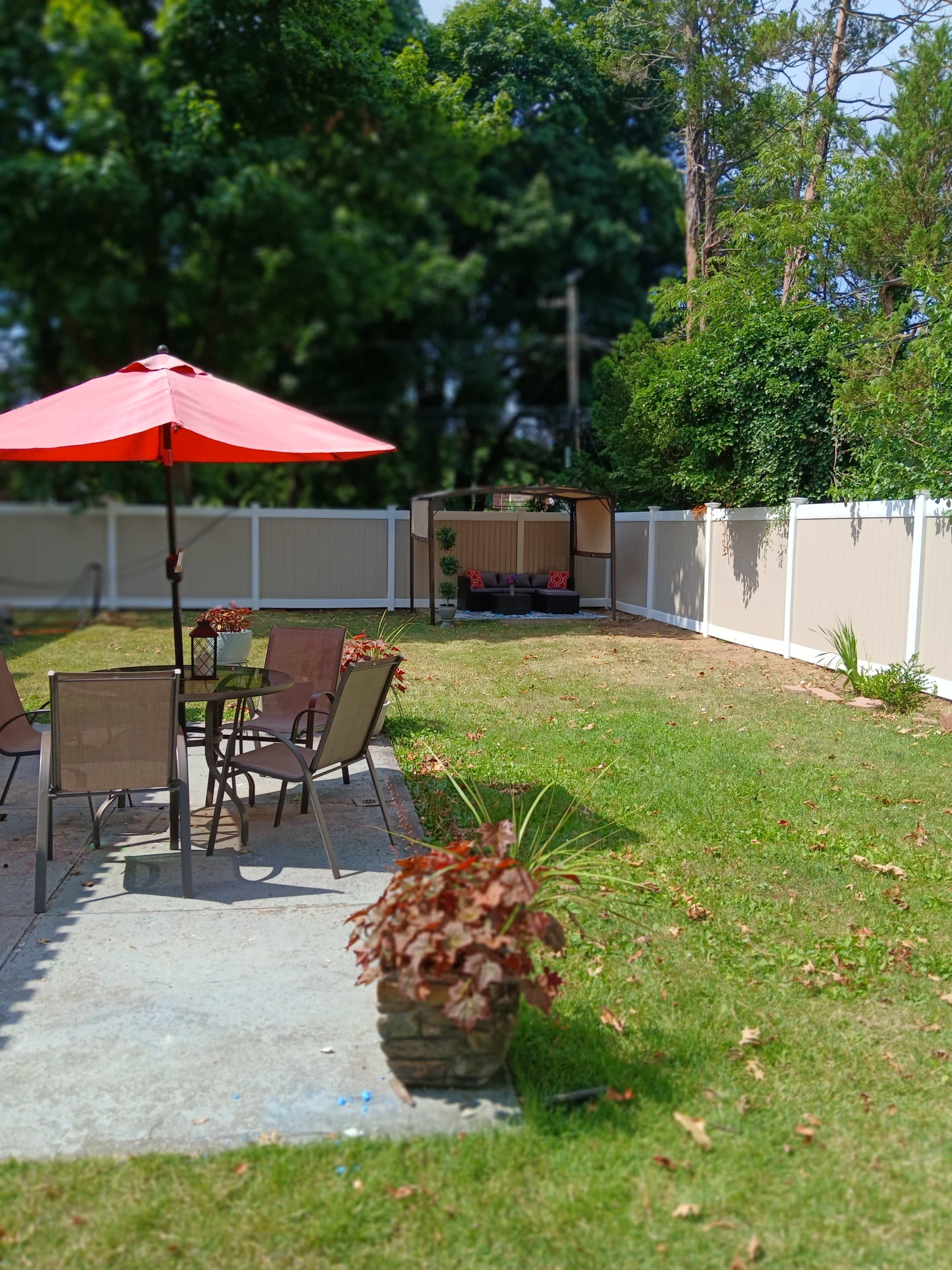 A backyard with a patio table and chairs under a red umbrella, surrounded by a fenced area and a storage shed in the background.