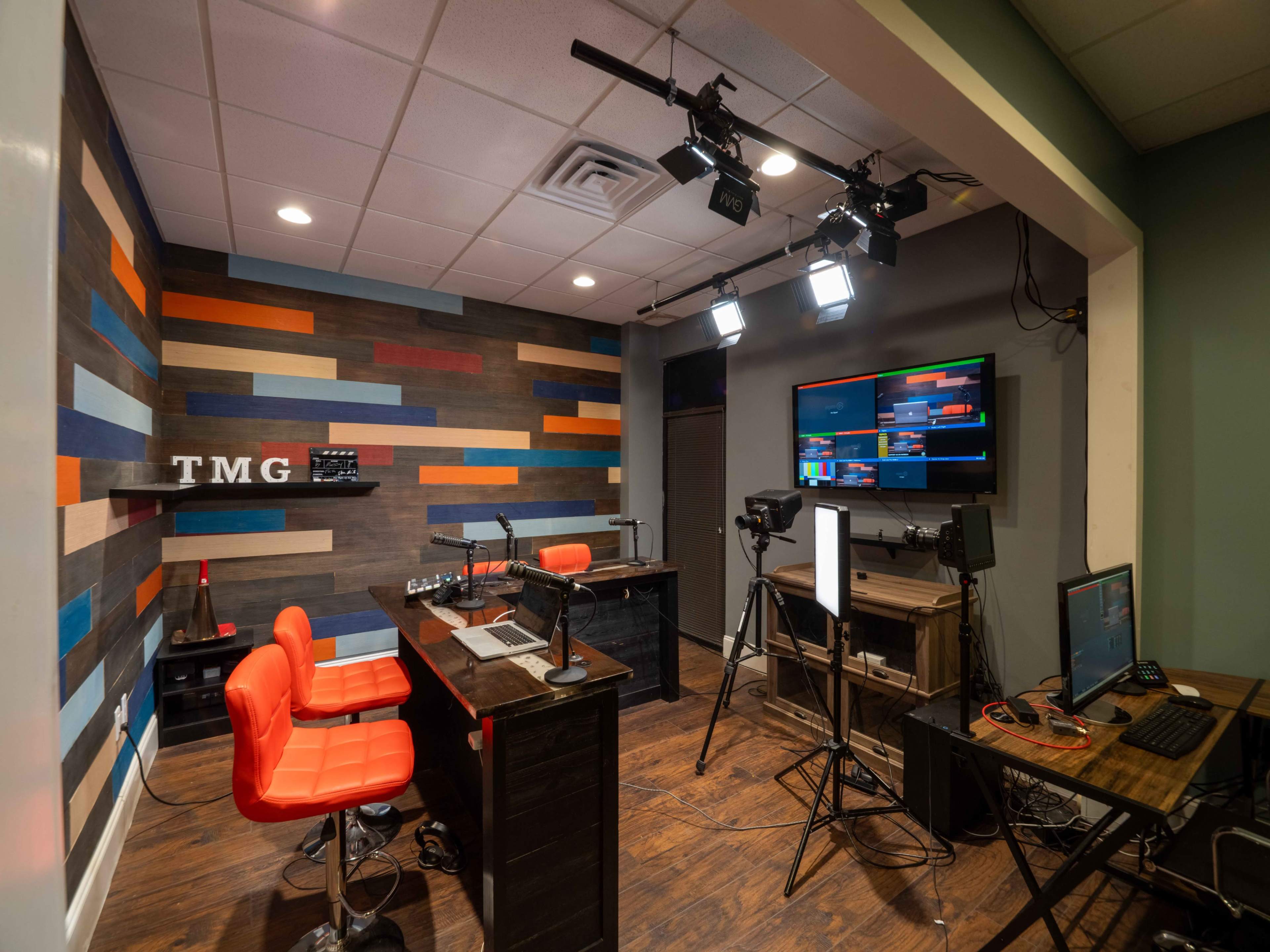 A studio setup with a wooden wall featuring colorful stripes, two orange chairs at a desk, and equipment for audio and video broadcasting.