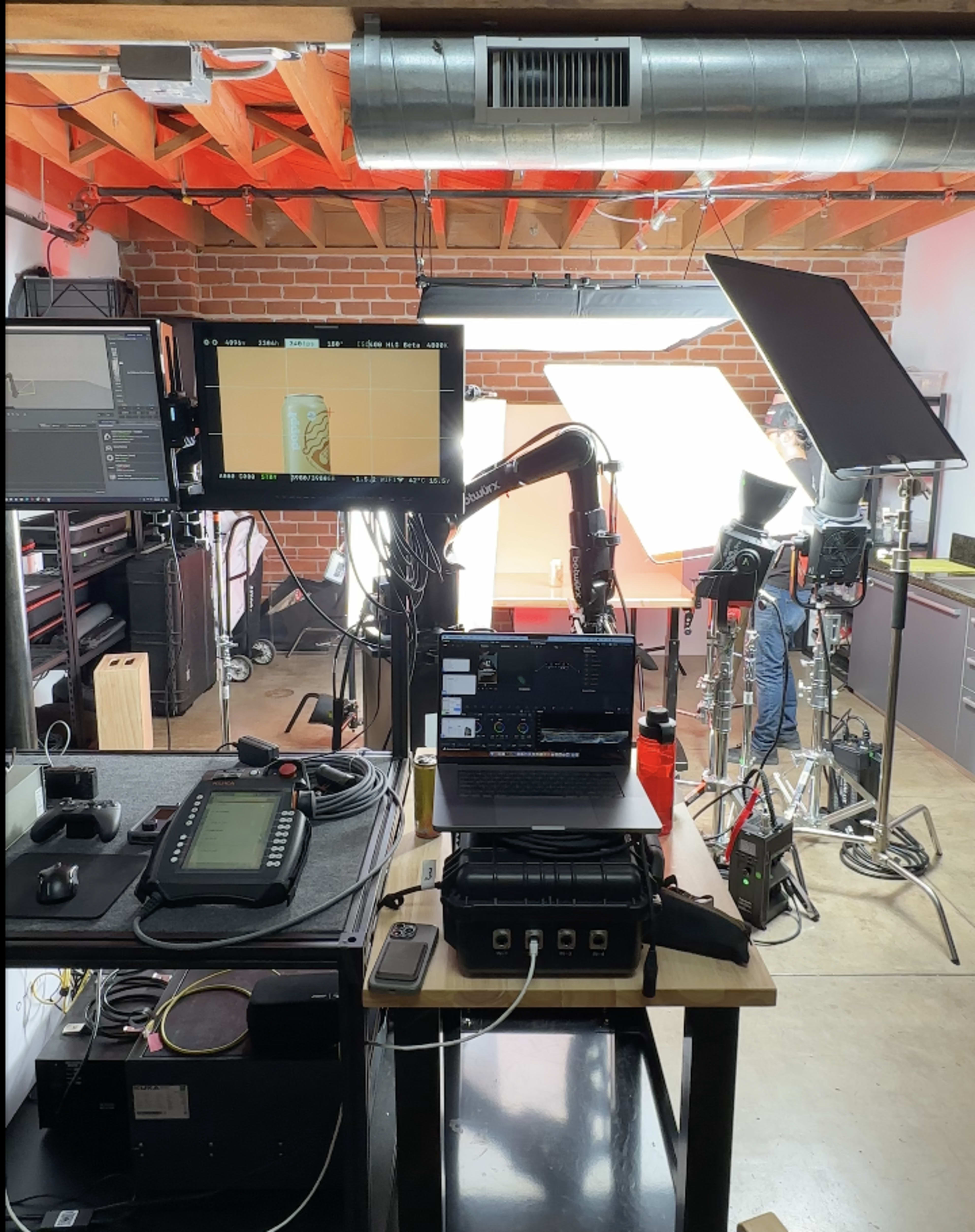 A photography studio setup features a laptop, cameras, and lighting equipment arranged on tables with exposed brick and wooden beams in the background.