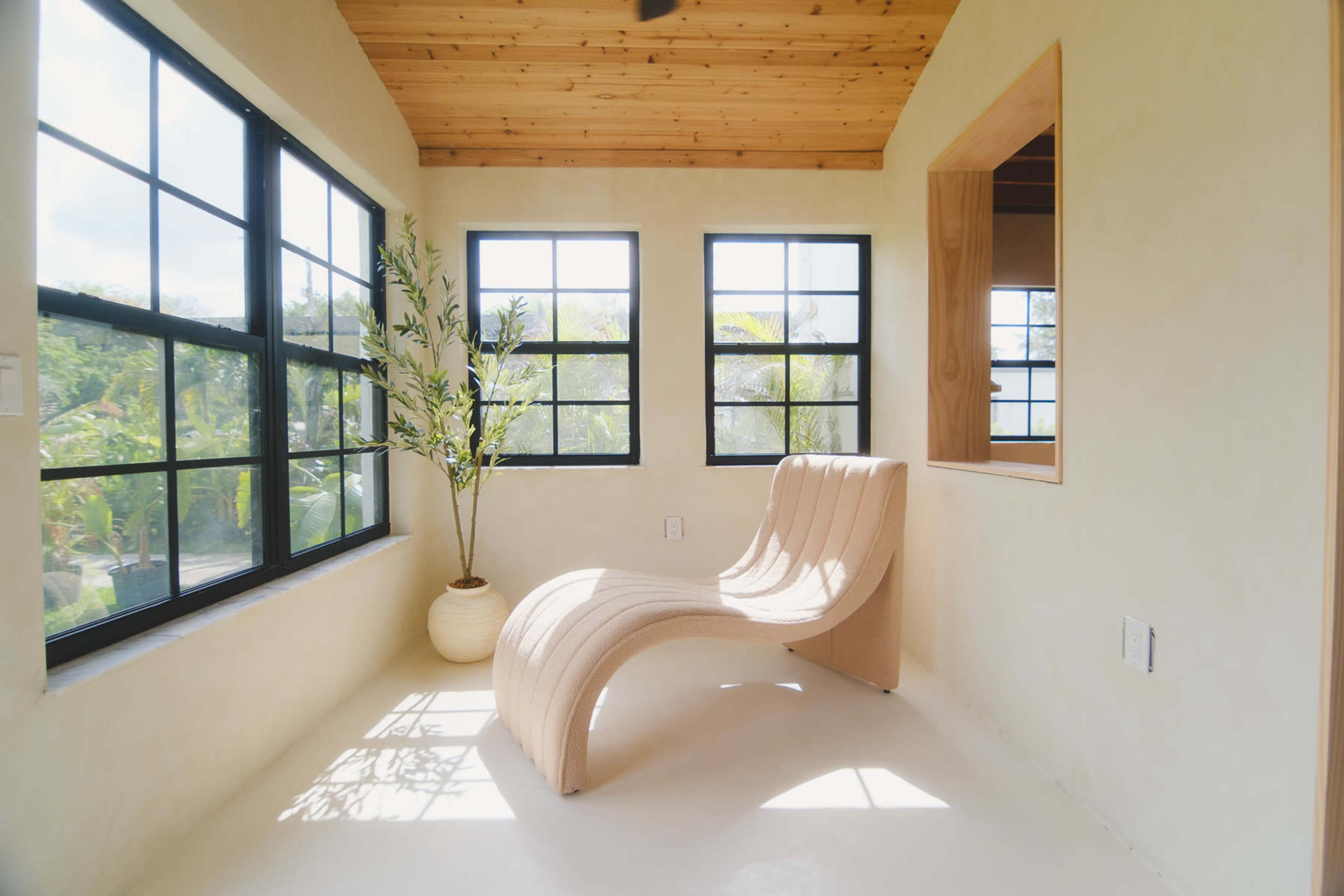 The image depicts a bright, minimalist room featuring a curved chair, large black-framed windows, and a potted plant.