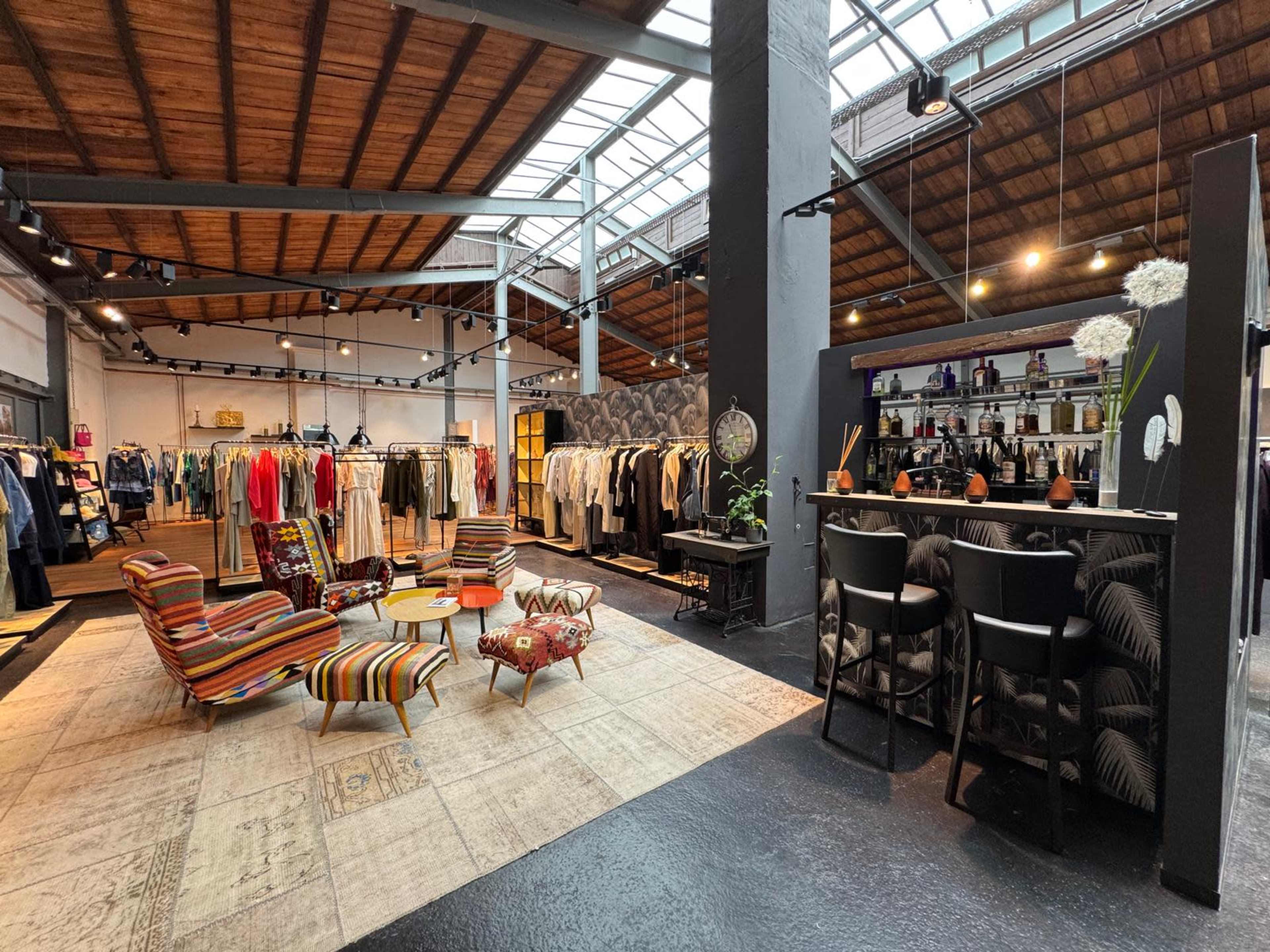 The image shows a stylish clothing store featuring a central seating area with colorful chairs and a bar displaying fashion items along one wall.