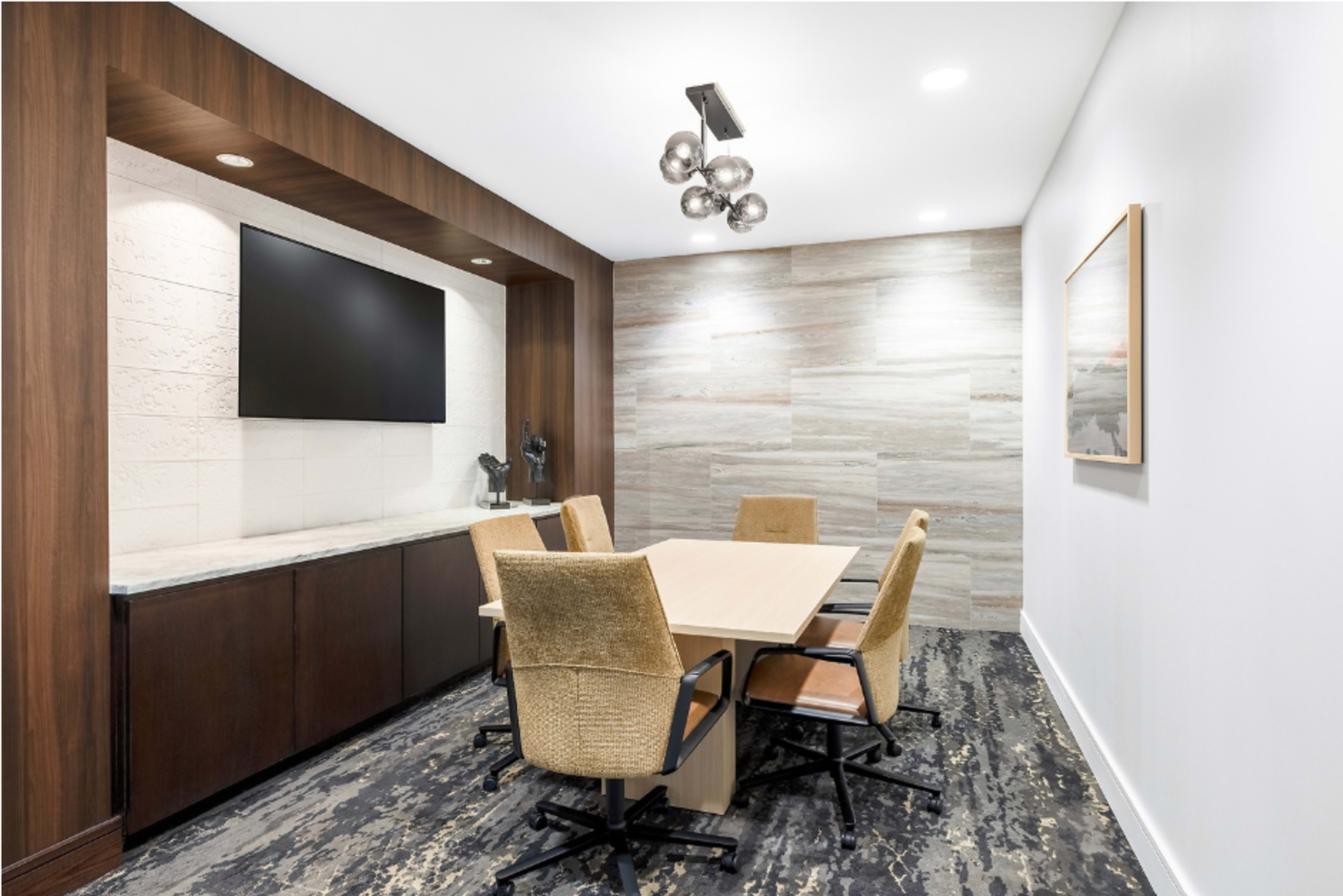 A conference room features a rectangular table surrounded by four chairs, with a large wall-mounted television and decorative wall paneling.