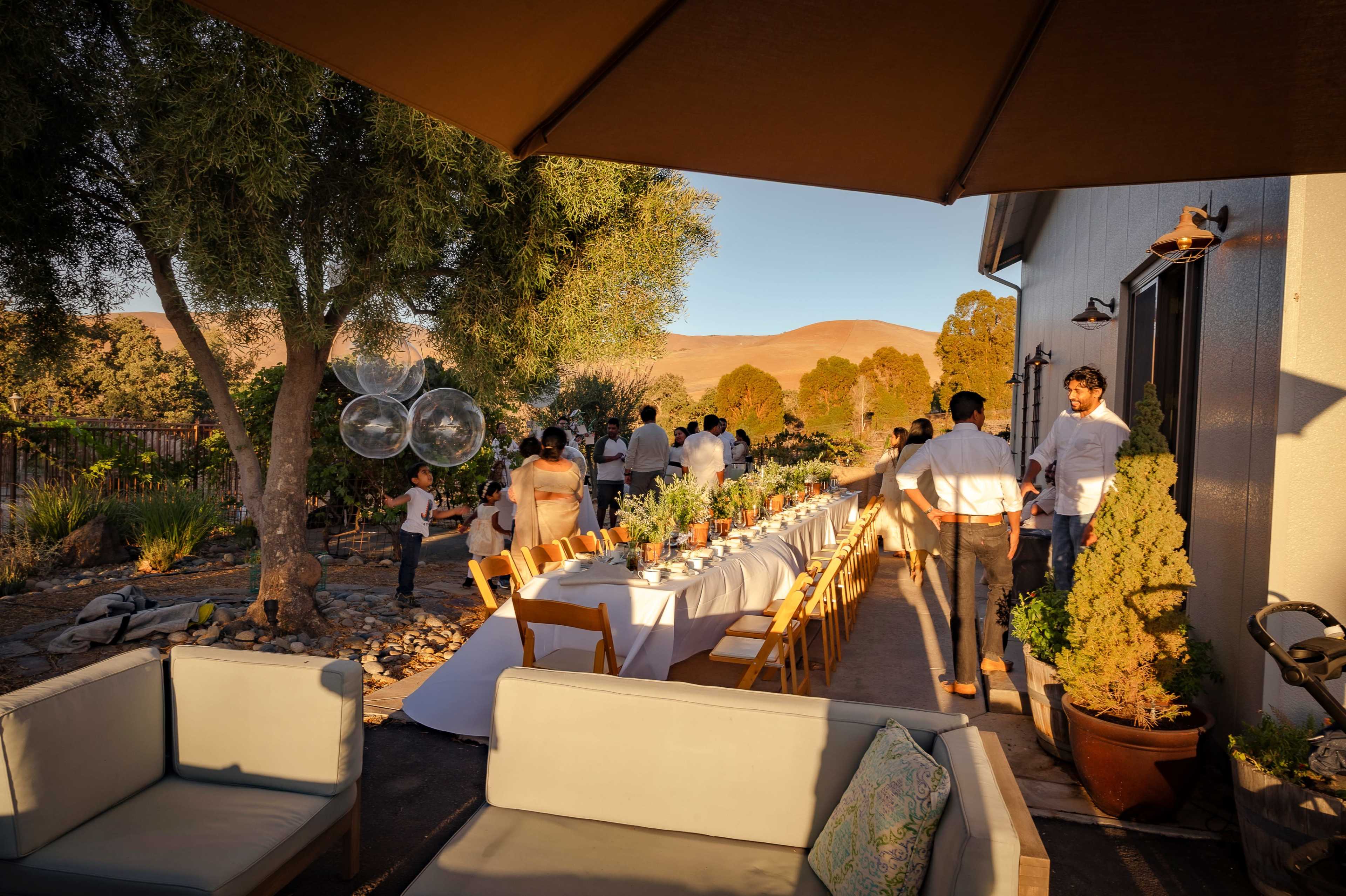 A long dining table is set up outdoors under an umbrella, surrounded by guests and greenery, with a mountainous backdrop in the distance.