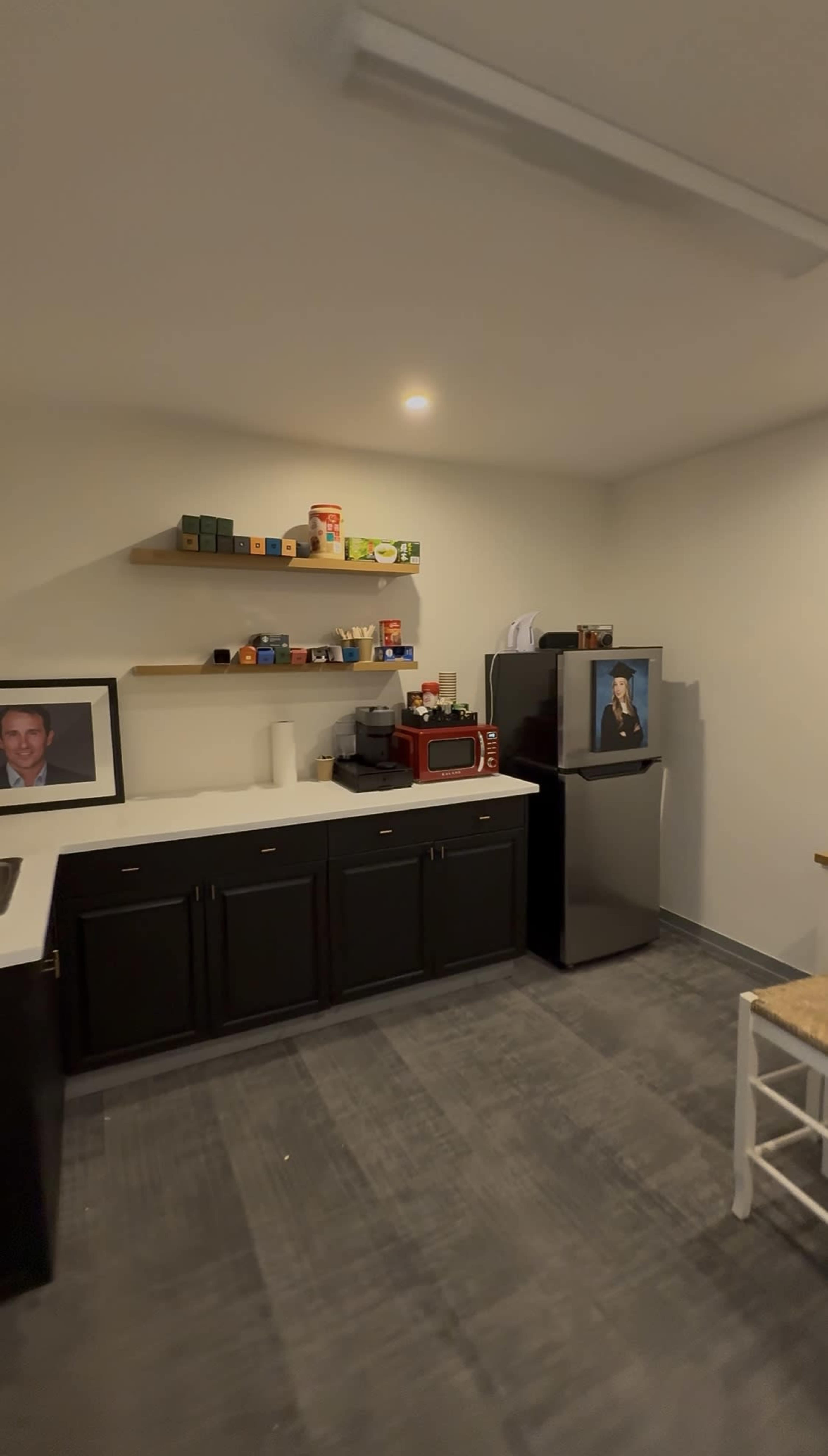 The image shows a kitchenette area with dark cabinets, a small red microwave, shelves filled with containers, and a refrigerator in the corner.
