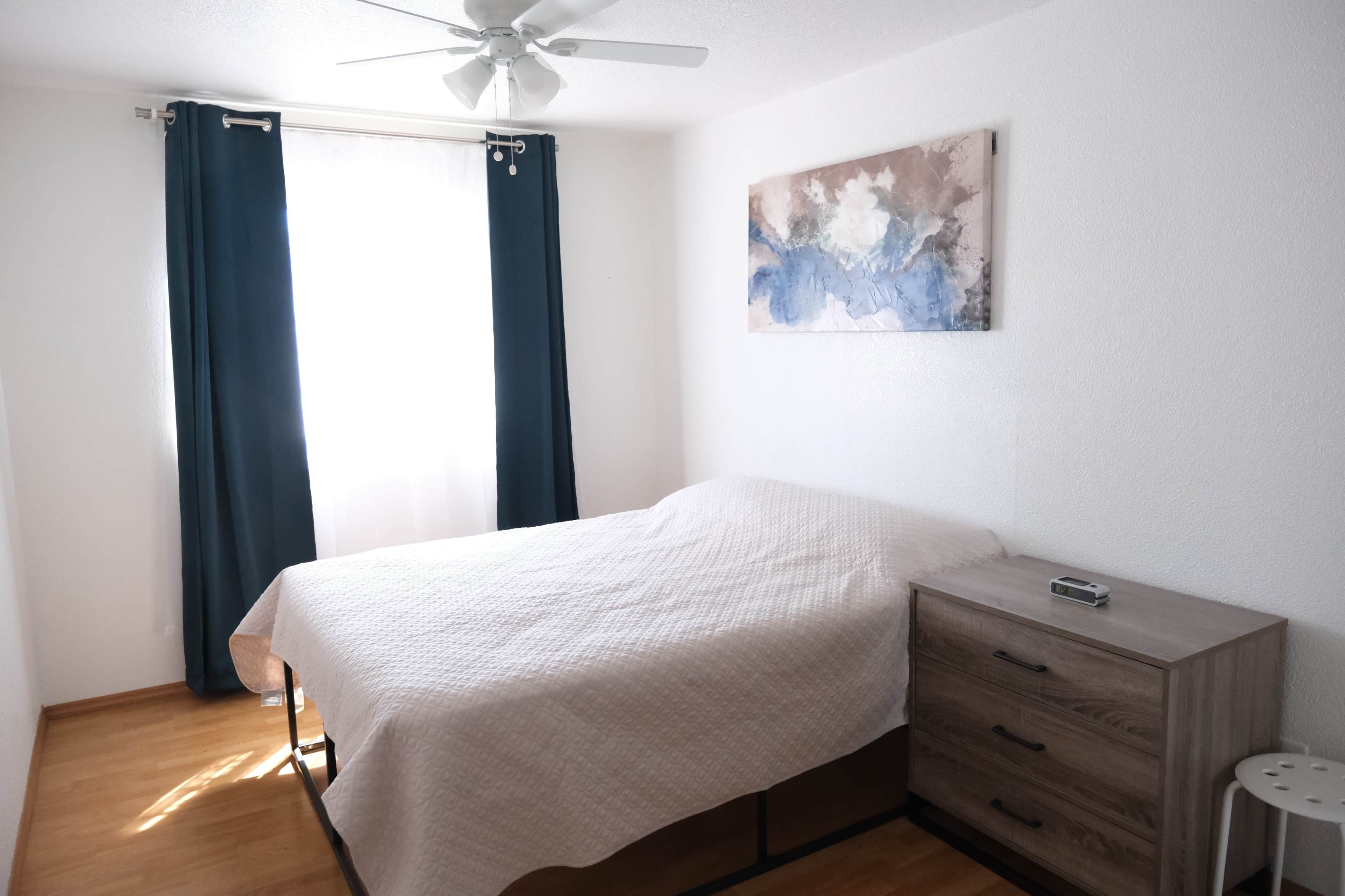 The image shows a simple bedroom with a bed covered by a quilt, a wooden nightstand, a window with curtains, and a wall-mounted painting.