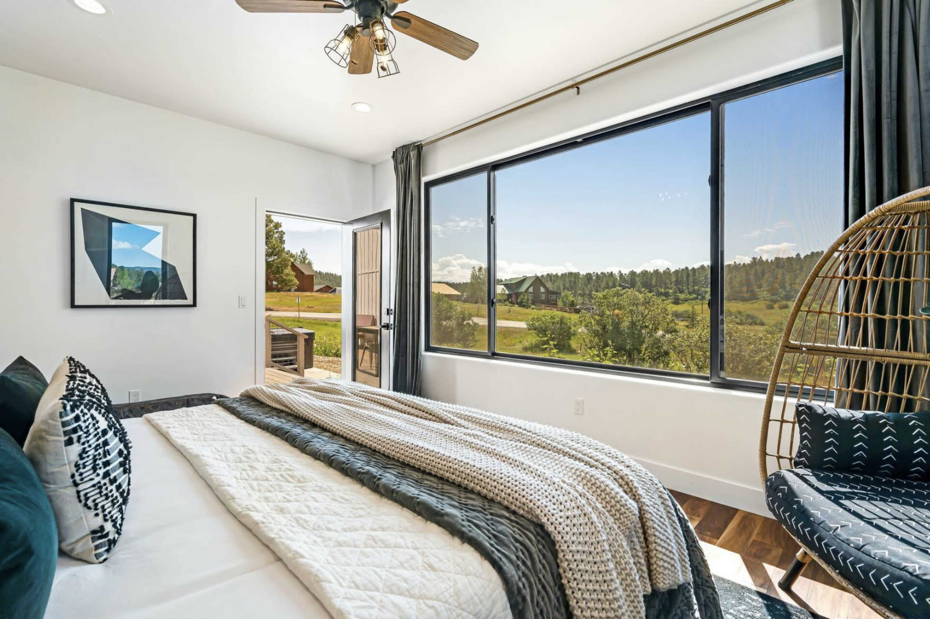 A modern bedroom features a large window overlooking a scenic view, with a bed draped in a quilt and a stylish hanging chair in the corner.