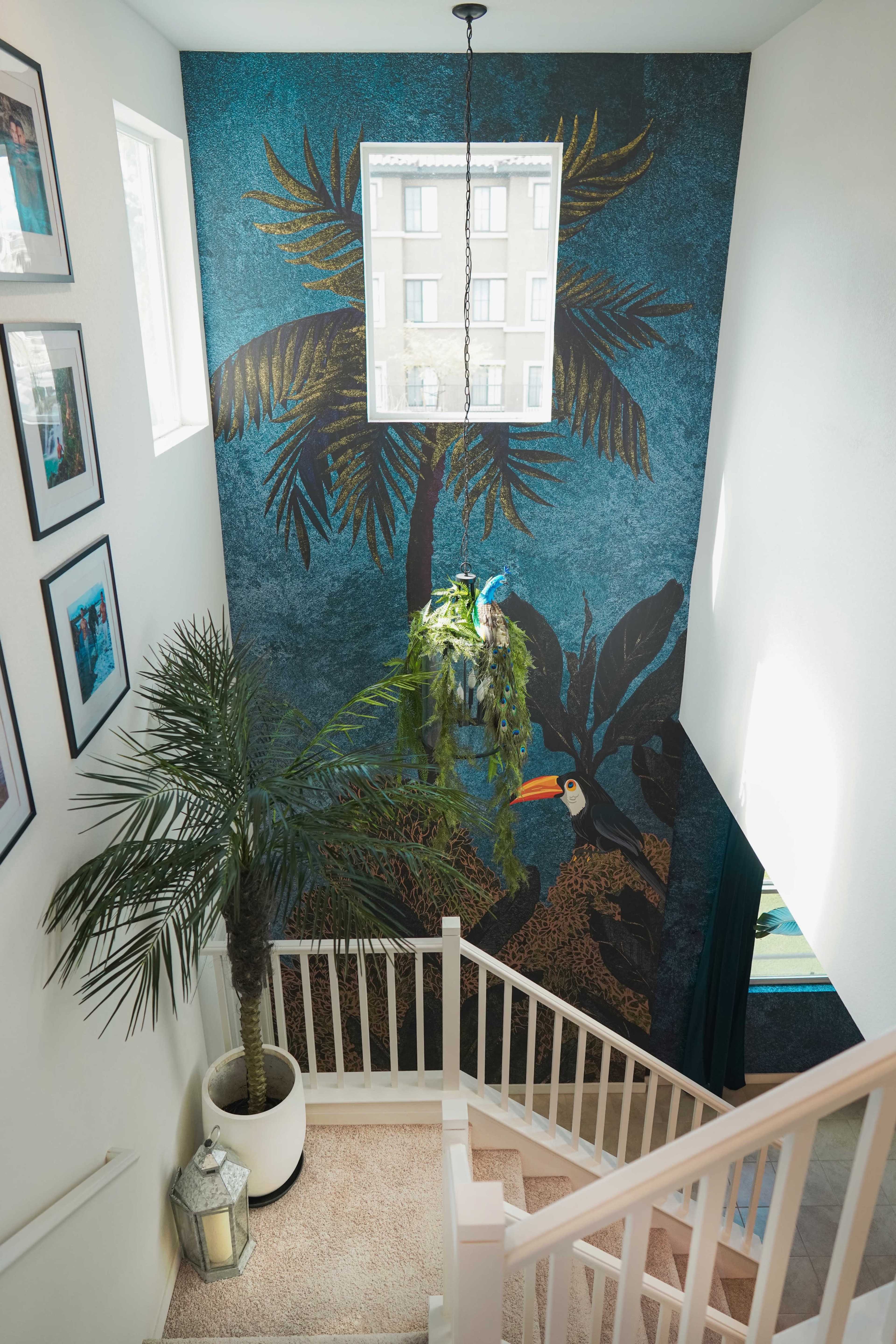 A staircase with a tropical mural on the wall, framed photos on the sides, and a potted palm at the bottom.