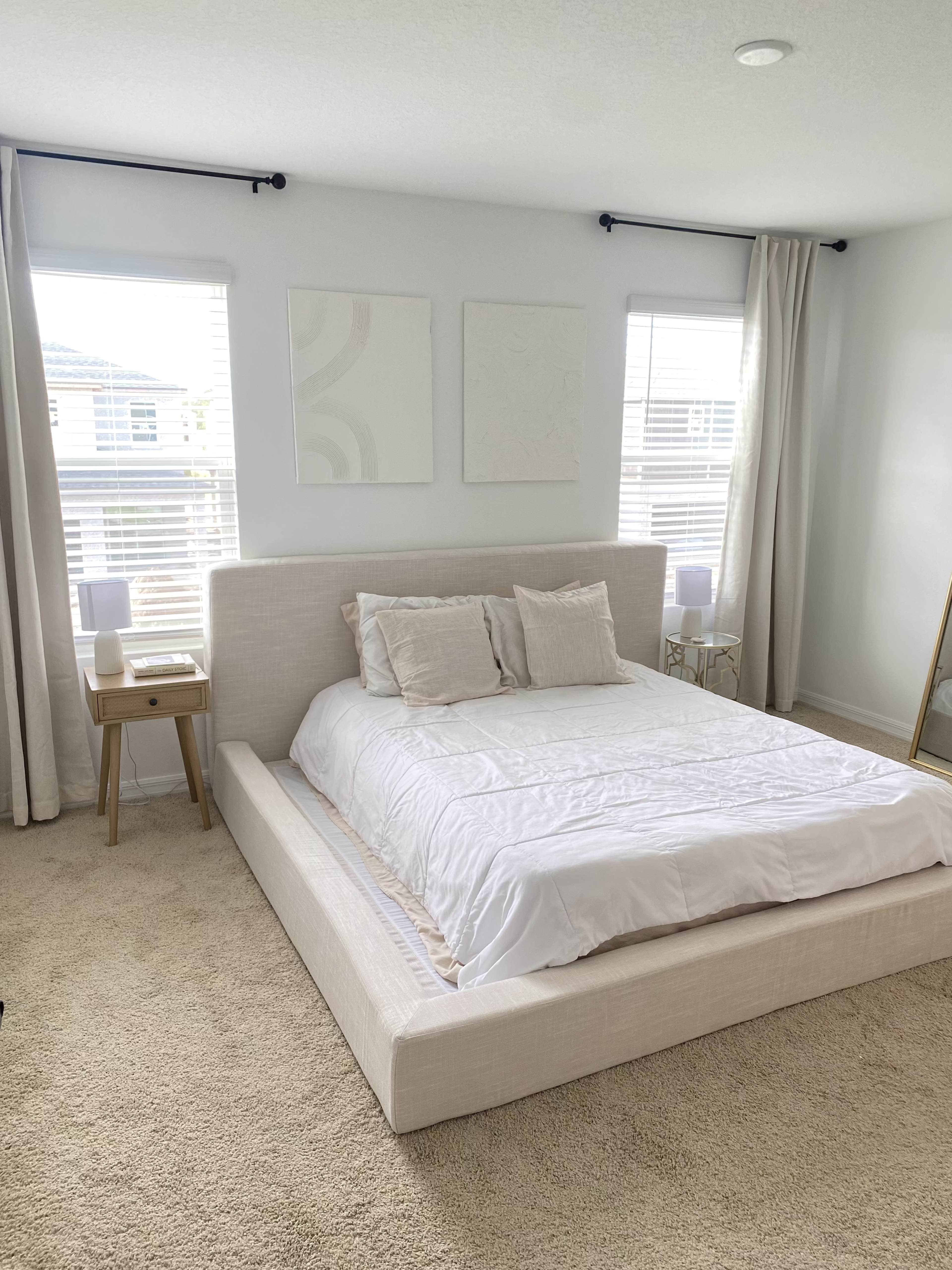 A neatly arranged bedroom features a low-profile bed with white bedding, two bedside lamps, and minimal artwork on the walls, illuminated by natural light from two windows.