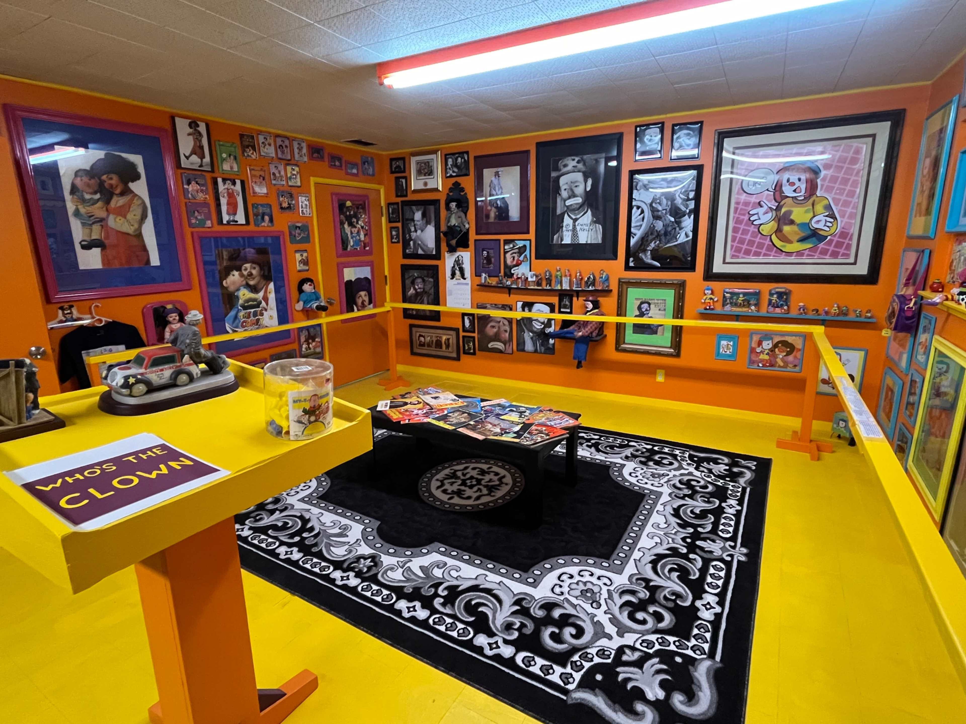 The image depicts a vibrant room filled with colorful walls adorned with various framed pictures and memorabilia, including a central coffee table covered with magazines and toys.