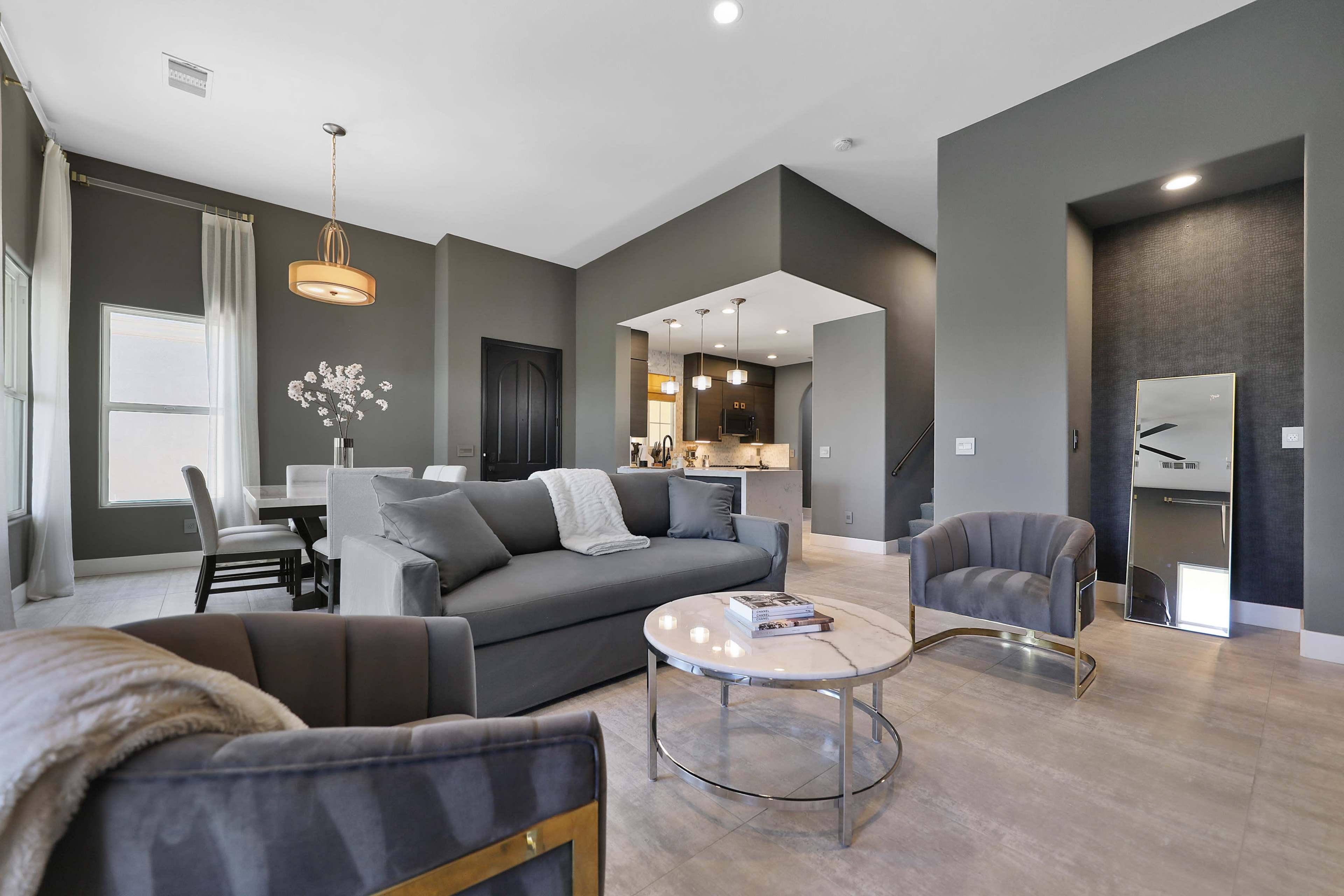 The image shows a modern interior living space featuring a gray sofa, two armchairs, a round coffee table, and a dining area visible in the background.