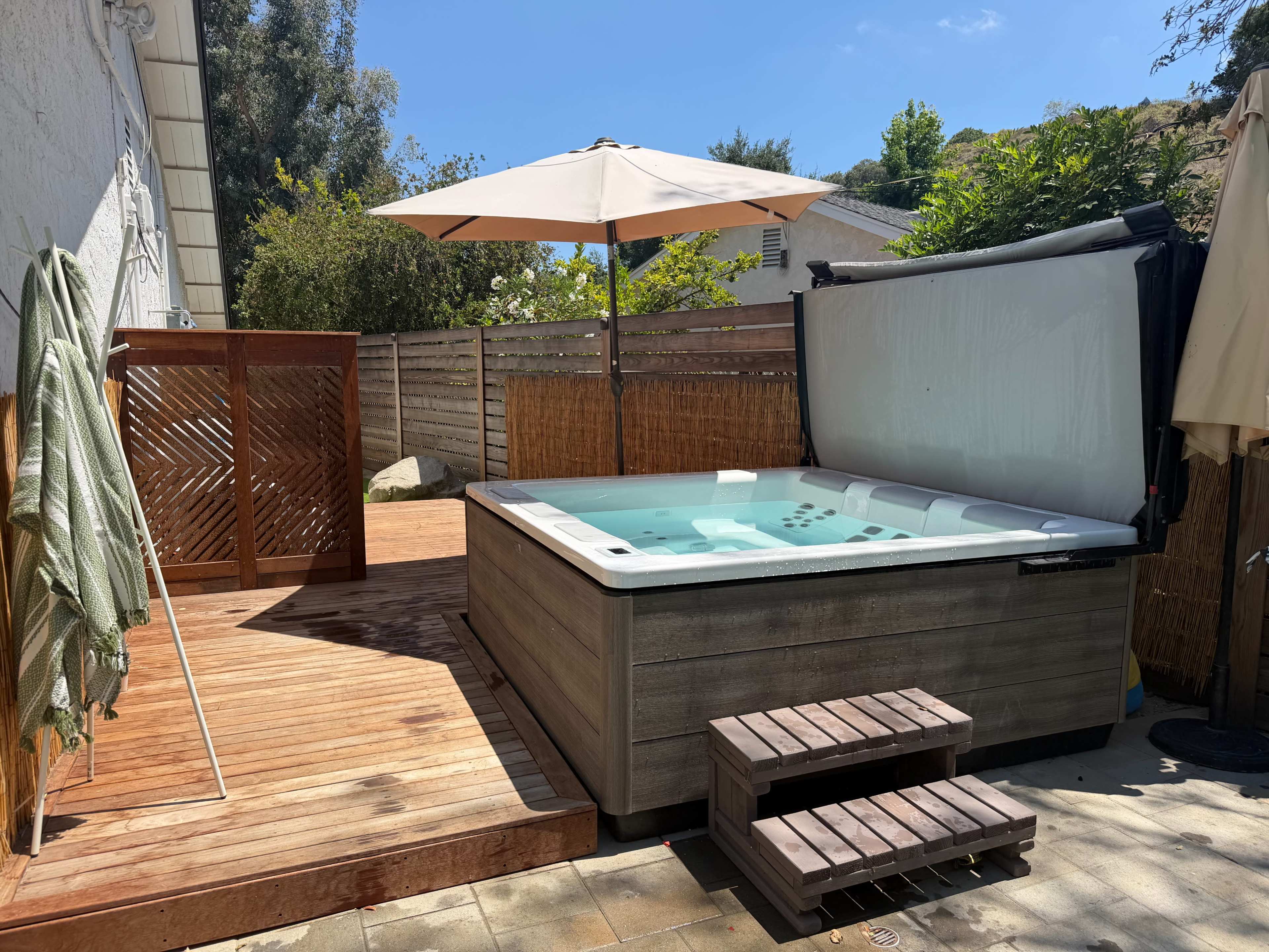 A hot tub sits on a wooden deck under a sun umbrella in a backyard surrounded by greenery and a wooden fence.